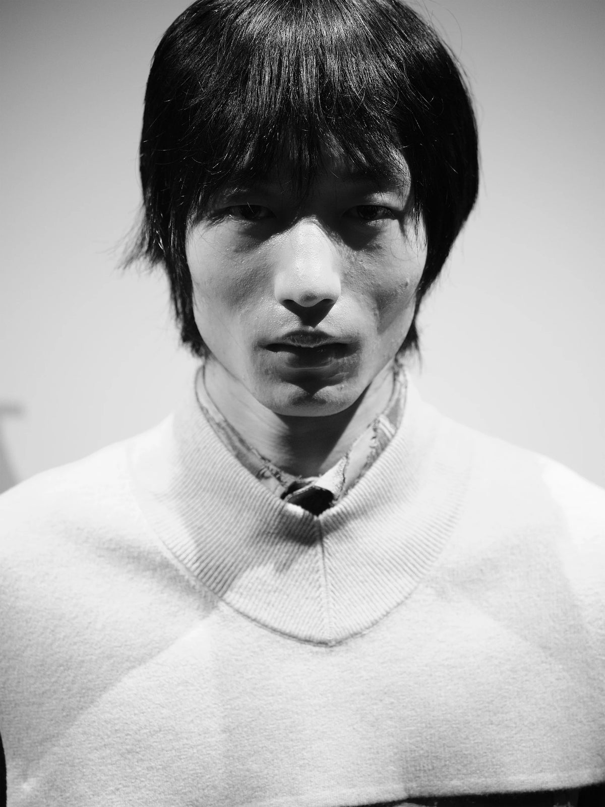 Black and white close-up portrait of a male model with a straight dark fringe, wearing a cream ribbed V-neck wool sweater layered over a collared shirt with a raw-edged denim neckline detail and a thin chain necklace, against a pale grey background. AW26 Icicle Atelier Menswear.