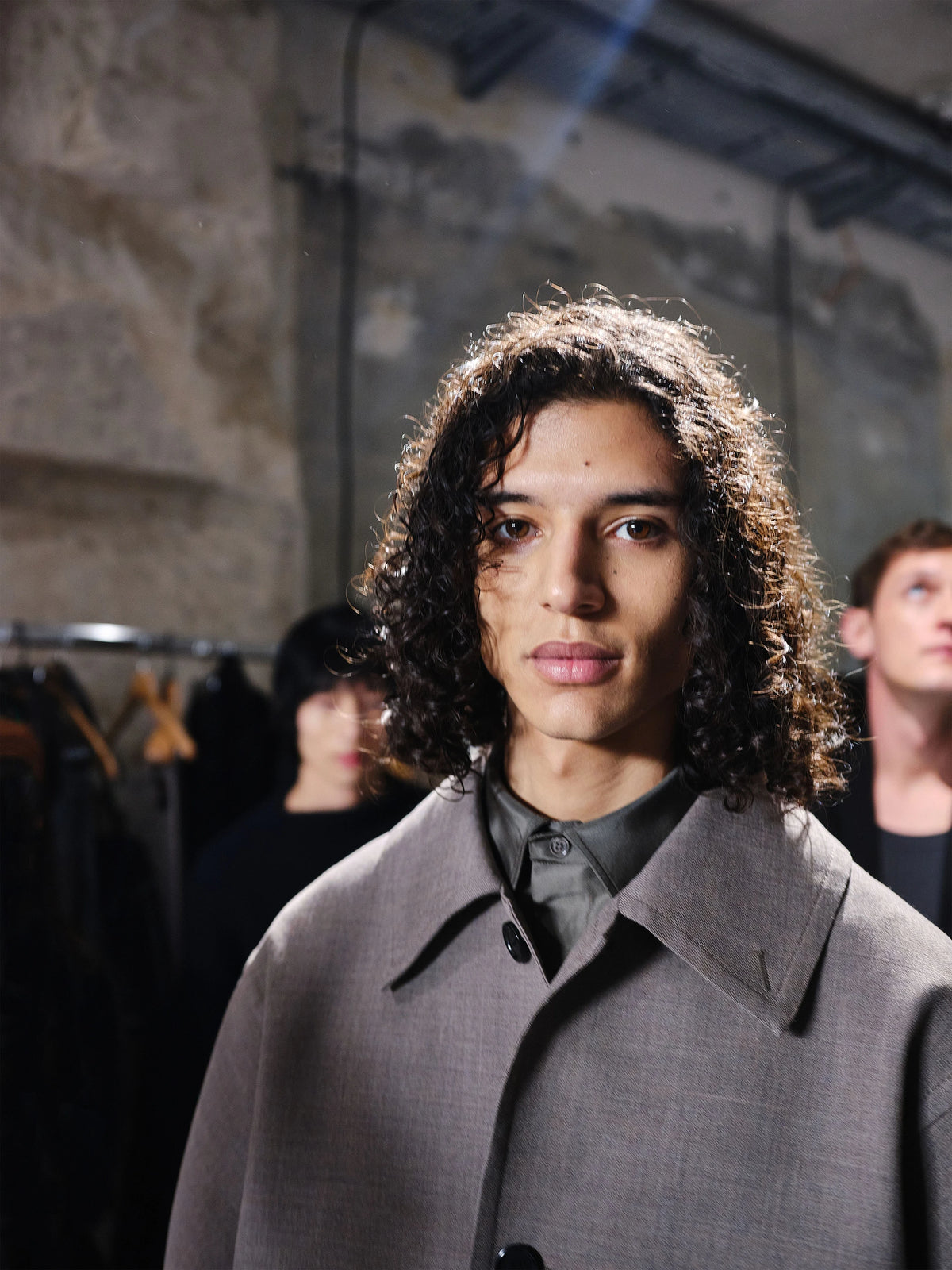 Backstage close-up portrait of a male model with long curly dark brown hair with blonde highlights, wearing an oversized light grey wool collarless coat with a single black button, layered over a dark grey collared shirt, in a dimly lit stone-walled backstage space with clothing rails and other models visible behind. AW26 Icicle Atelier Menswear.