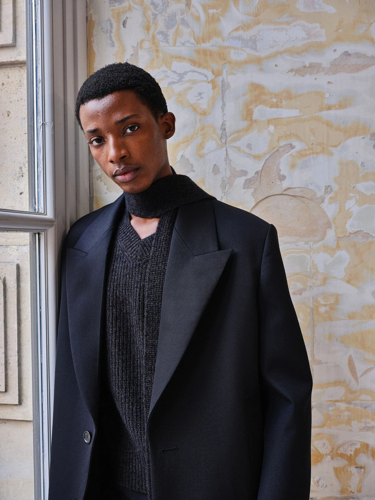 Male model leaning against a weathered Parisian stone wall beside a tall window, wearing an oversized black wool double-breasted blazer with wide peaked lapels and a single horn button, layered over a dark charcoal grey ribbed V-neck knit with a black turtleneck underneath. AW26 Icicle Atelier Menswear.