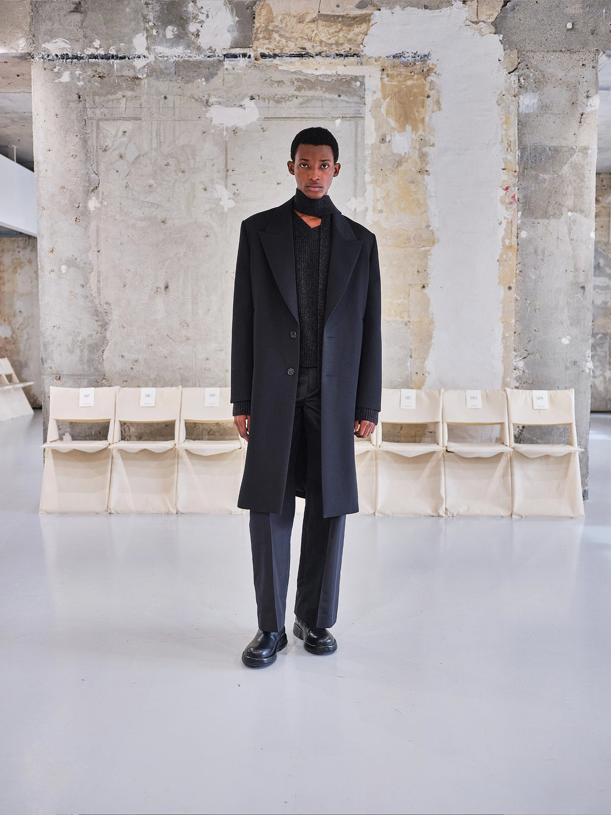 Male model standing on a white runway floor in a raw Parisian stone venue with cream folding chairs behind him, wearing a full-length black wool single-breasted overcoat with wide peaked lapels and two buttons, layered over a dark charcoal ribbed knit and black turtleneck, paired with wide-leg dark grey trousers and black chunky leather loafers. AW26 Icicle Atelier Menswear.