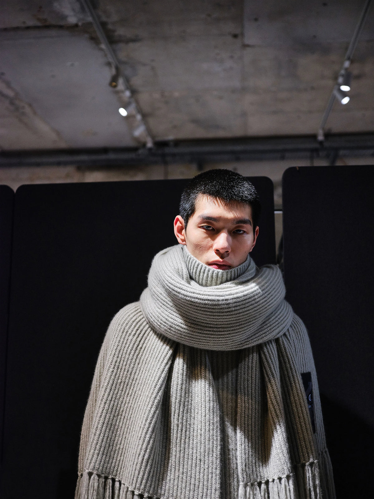 Backstage close-up portrait of a male model with short dark hair, wearing an oversized light grey marl chunky ribbed-knit turtleneck sweater with an integrated extra-long scarf wrapped voluminously around the neck and a small woven label badge on the chest, under dramatic spotlight lighting against a dark background. AW26 Icicle Atelier Menswear.