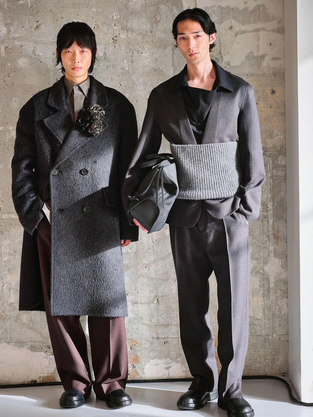Two male models standing against a raw concrete wall: left model wears a knee-length double-breasted dark grey textured bouclé wool coat with wide lapels, a black sculptural floral brooch, a khaki shirt underneath, dark brown wide-leg trousers and black chunky loafers, holding a grey leather saddle bag; right model wears a dark grey silk jacquard open-collar shirt with a wide grey ribbed knit cummerbund waistband, matching wide-leg grey trousers and black chunky loafers. AW26 Icicle Atelier Menswear.