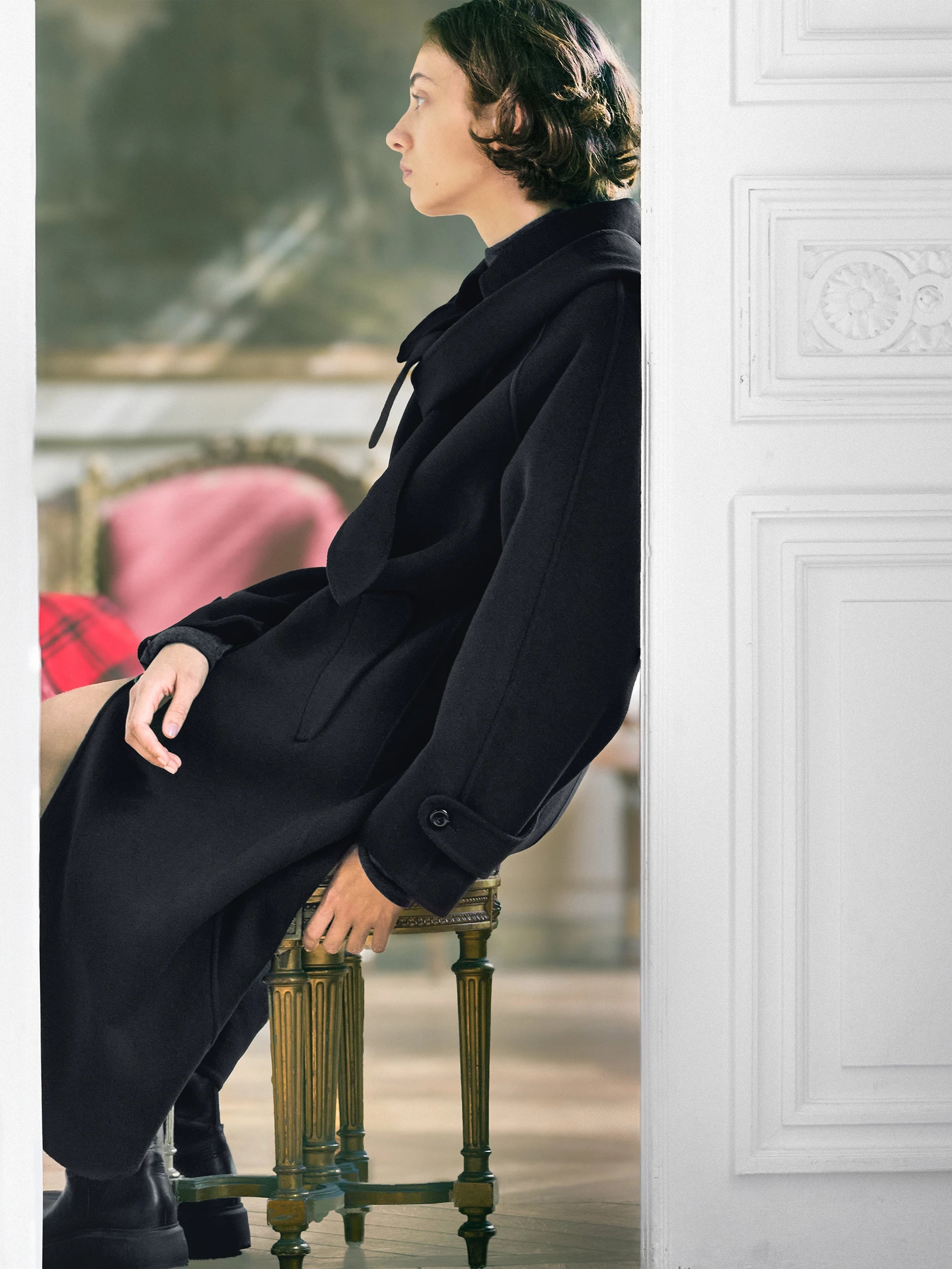 A woman wearing a long black coat sits on a wooden stool in an elegant interior setting, framed by a white paneled door on the right.