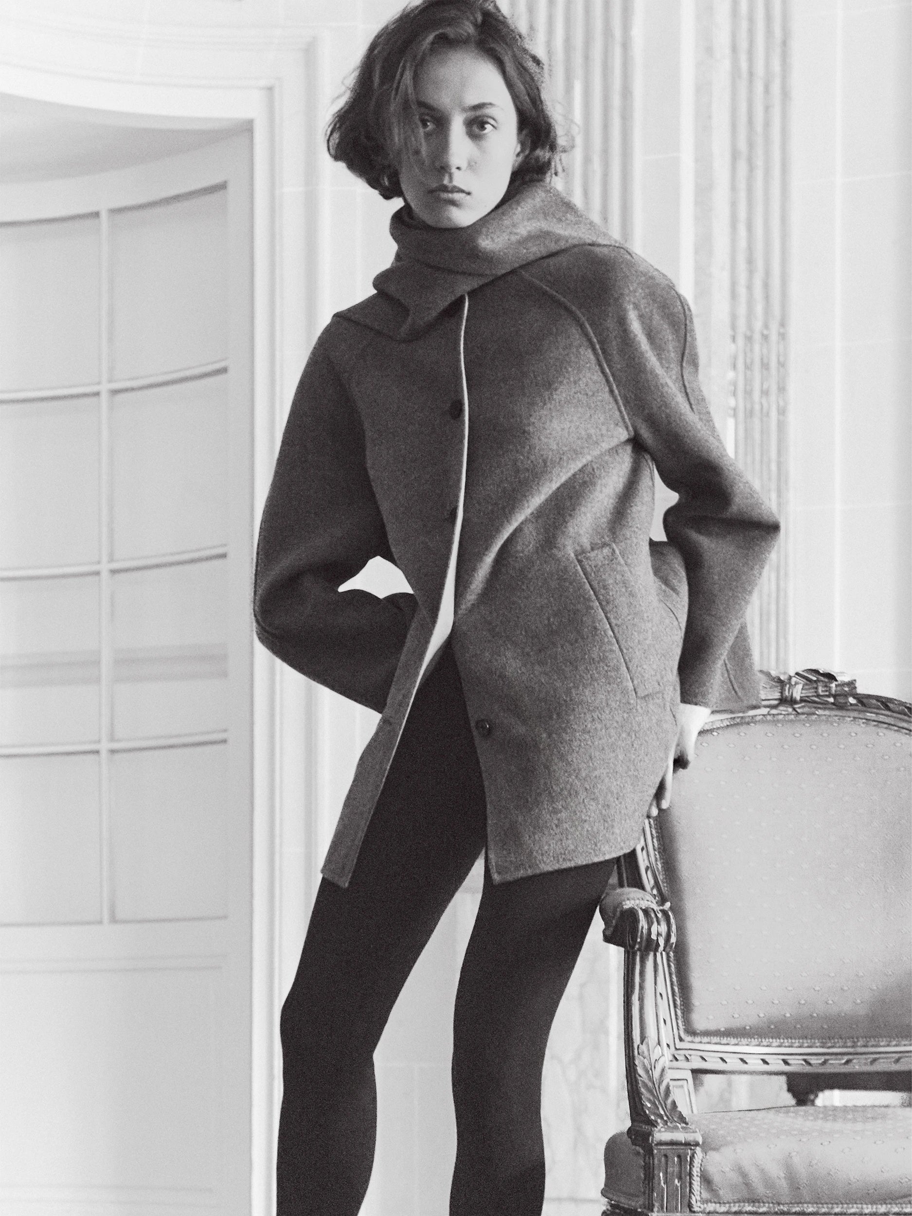 A woman wearing a gray oversized coat with a high collar and dark tights, standing indoors next to an ornate chair in a bright, light-colored room.