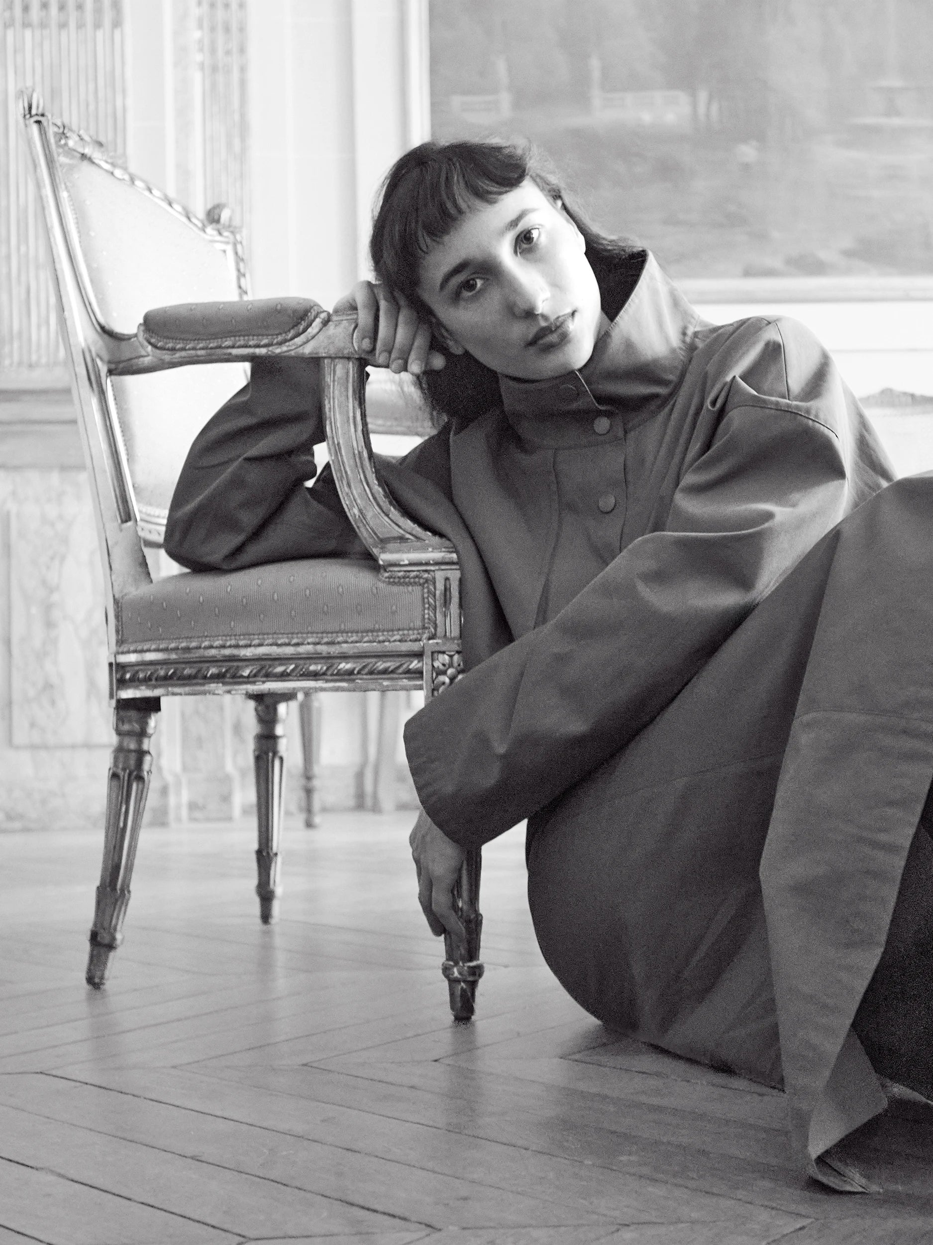 A black and white fashion photograph of a model wearing a dark oversized coat, leaning against an ornate vintage chair in a room with wooden floors.