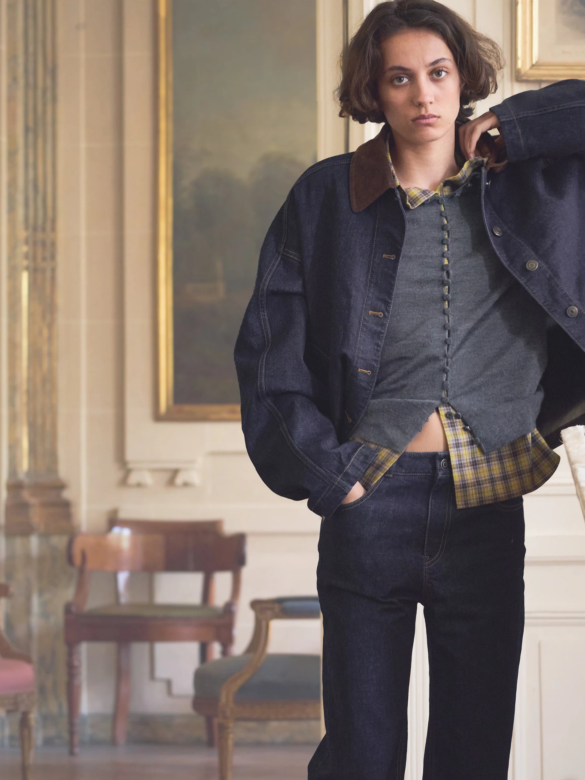 A model wearing a dark blue denim jacket with a brown corduroy collar, layered over a gray button-up shirt and dark blue jeans, standing in an elegant indoor room with classical paintings and wooden chairs.