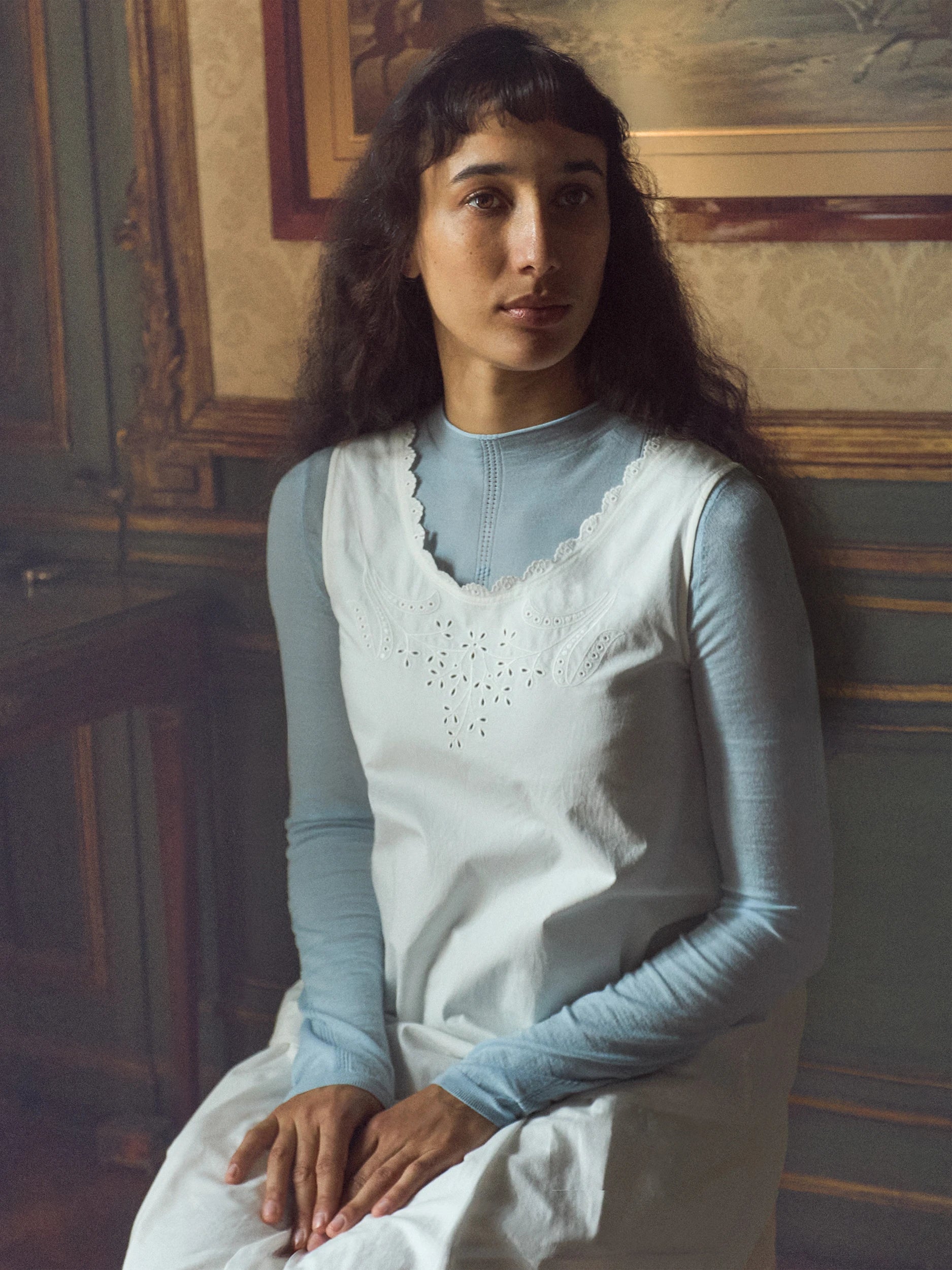 A woman seated indoors wearing a light blue long-sleeve top under a white eyelet dress or tunic with a scalloped neckline.