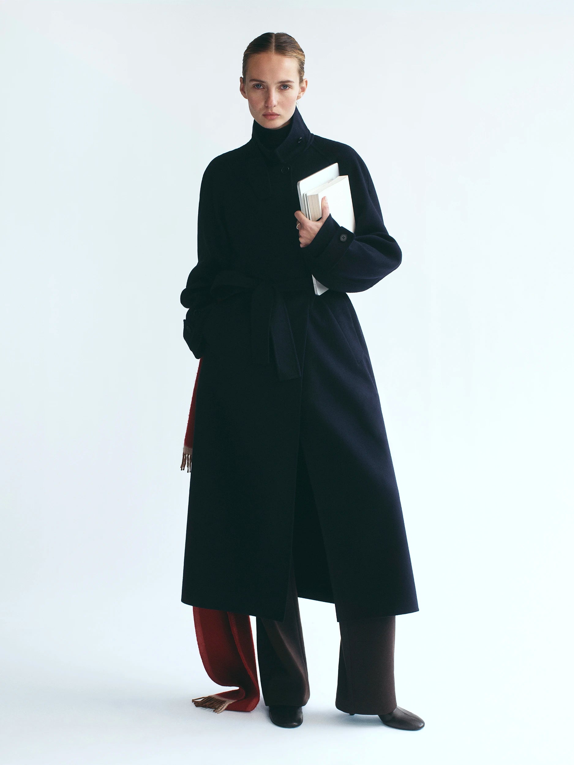 Model standing against a white studio background wearing a floor-length navy wool coat with a high turtleneck, wide A-line silhouette and side pockets, holding a white hardcover book, with dark brown wide-leg trousers, a burgundy fringed scarf draped at the side, and dark leather boots. AW26 D-Line.