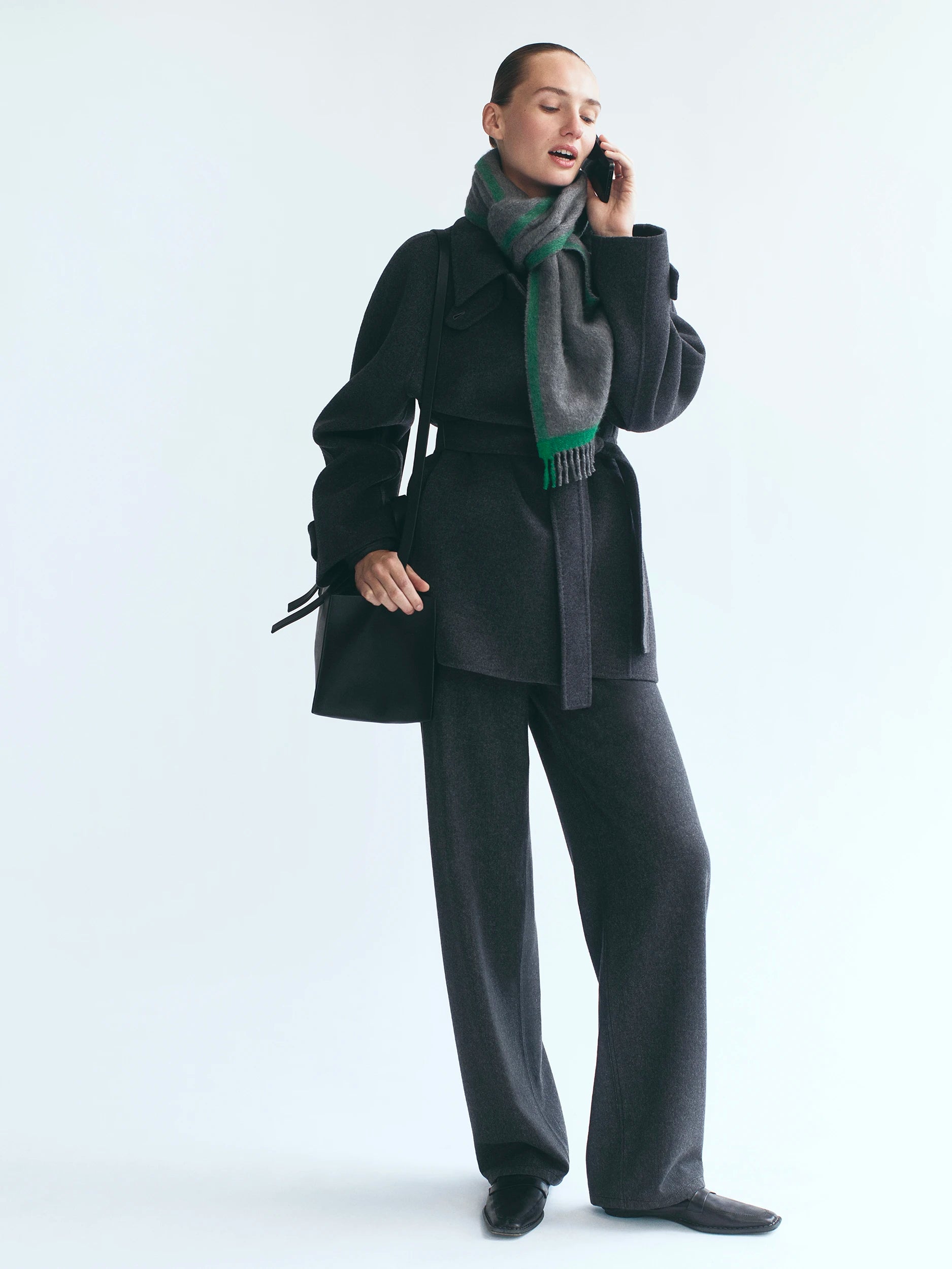 Model standing against a white studio background, talking on a phone, wearing an oversized black wool belted jacket layered over a dark charcoal wide-leg trouser suit, with a large grey and green fringed wool scarf wrapped high around the neck, and black loafers. AW26 D-Line.