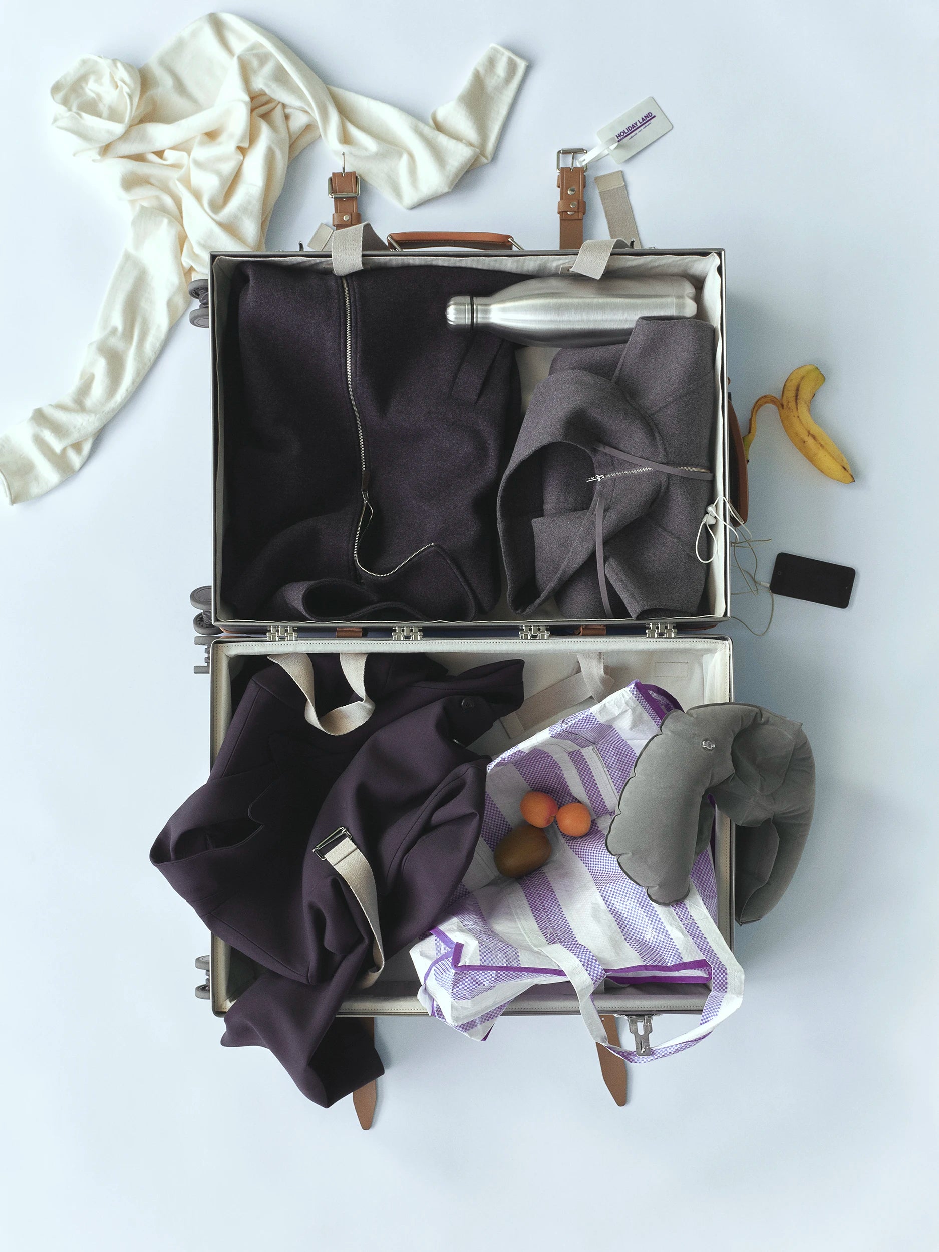Overhead flat-lay of an open hard-shell suitcase with tan leather trim, packed with dark navy and charcoal Icicle knitwear, a stainless steel water bottle and a grey travel pillow, alongside a striped tote bag with fruit, a cream draped garment and a banana peel scattered on a light grey surface. AW26 D-Line.