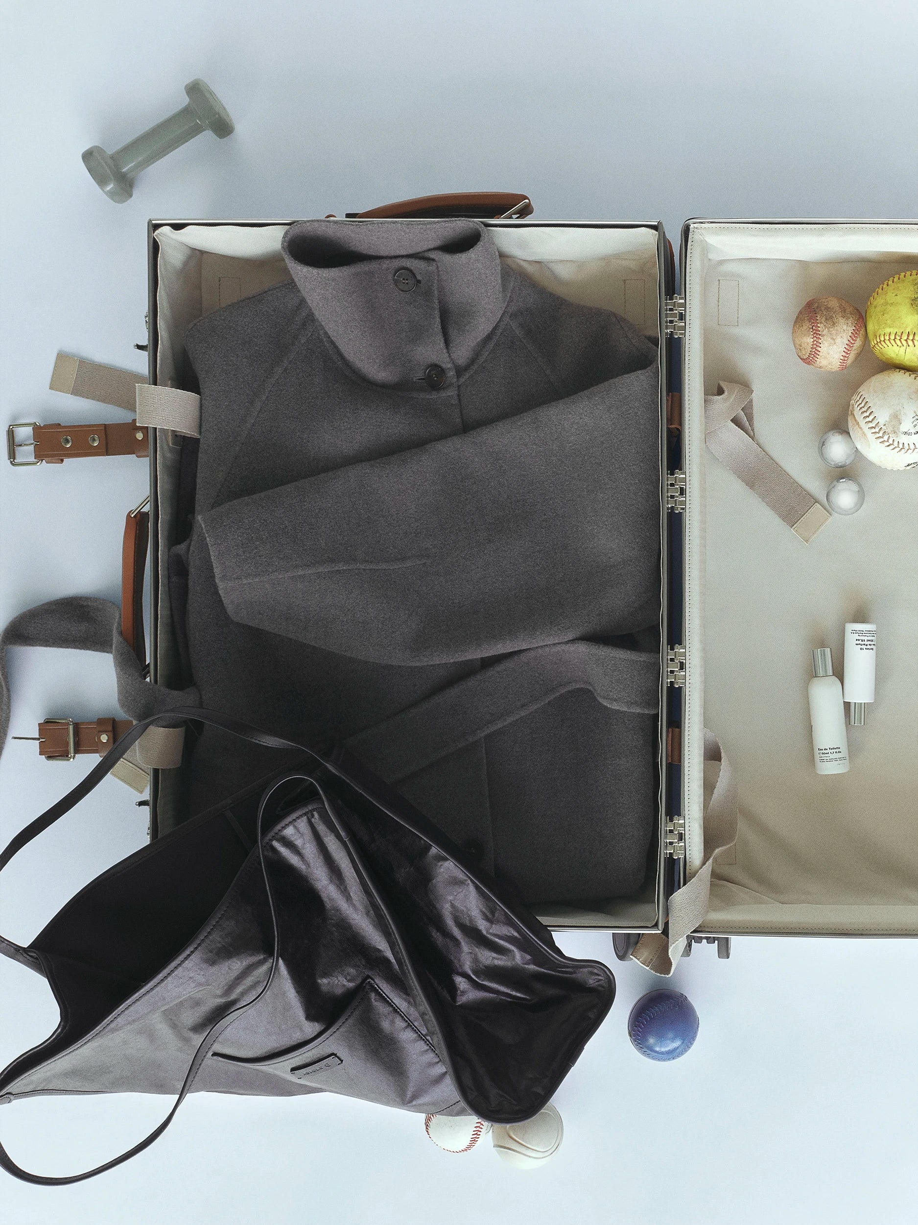 Overhead flat-lay of an open cream-lined suitcase with tan leather trim, packed with a folded dark charcoal wool coat with button collar detail; the right compartment holds baseballs, a yellow softball, pearl earrings and two white fragrance bottles; a black leather tote bag and a small dumbbell are placed beside the case. AW26 D-Line.