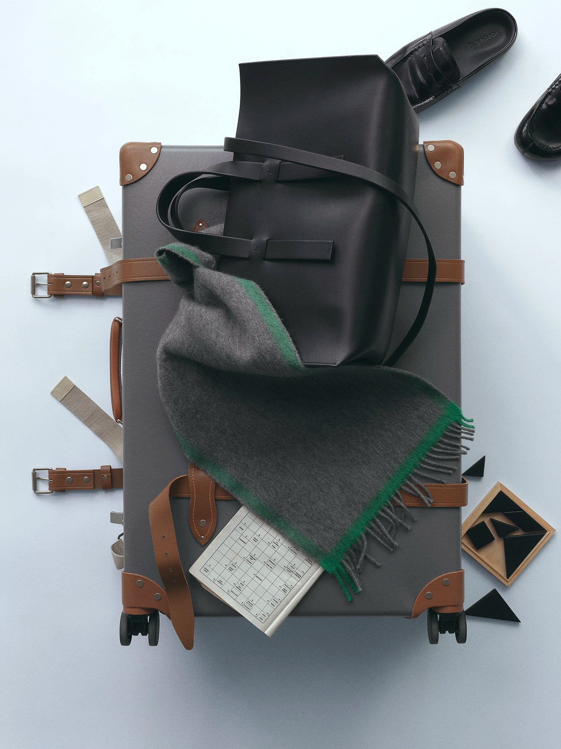Flat-lay overhead shot of a dark grey hard-shell trolley suitcase with tan leather trim, topped with a black leather tote bag, a grey and green fringed wool scarf, and a small notebook, with black Oxford shoes and accessories arranged around it. AW26 D-Line.