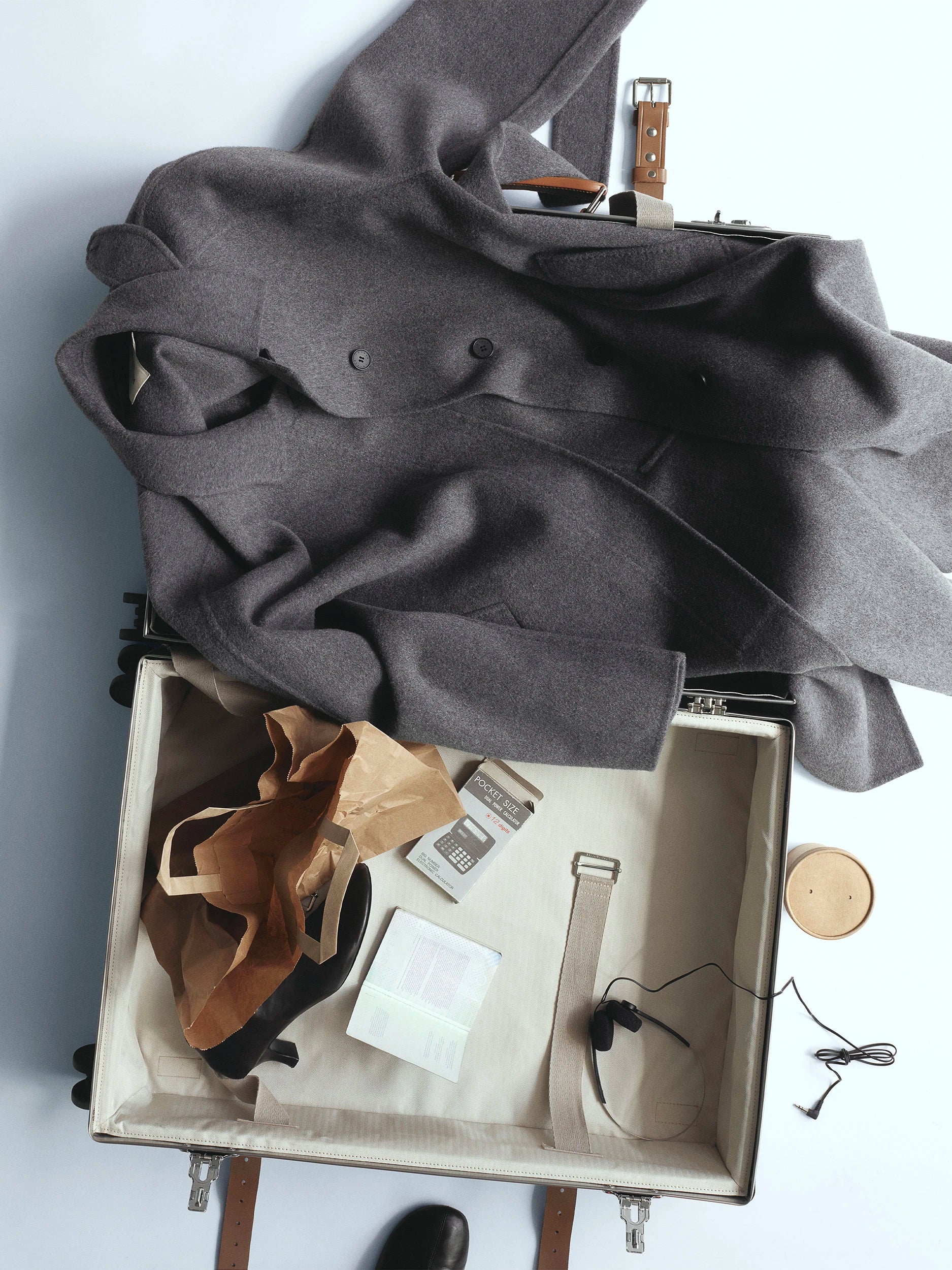 Open cream-lined suitcase with tan leather trim, the lid draped with a voluminous dark grey wool coat with double-button detail, the lower compartment holding black heeled sandals in a kraft paper bag, a pocket calculator, a notebook and earphones, with a wooden coaster and a black shoe visible beside it. AW26 D-Line.