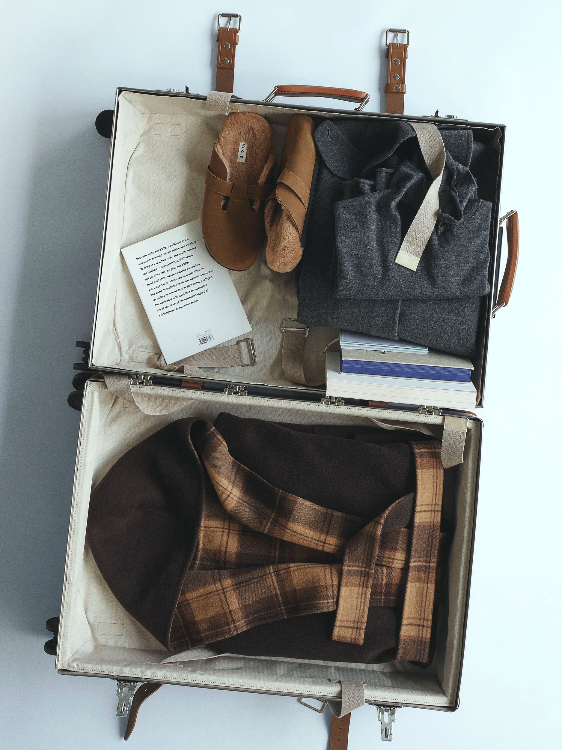 Open suitcase laid flat on a light grey surface, packed with Icicle pieces: tan leather sandals, a folded dark grey jacquard garment, stacked books, and a voluminous brown and camel tartan coat filling the lower compartment. AW26 D-Line.