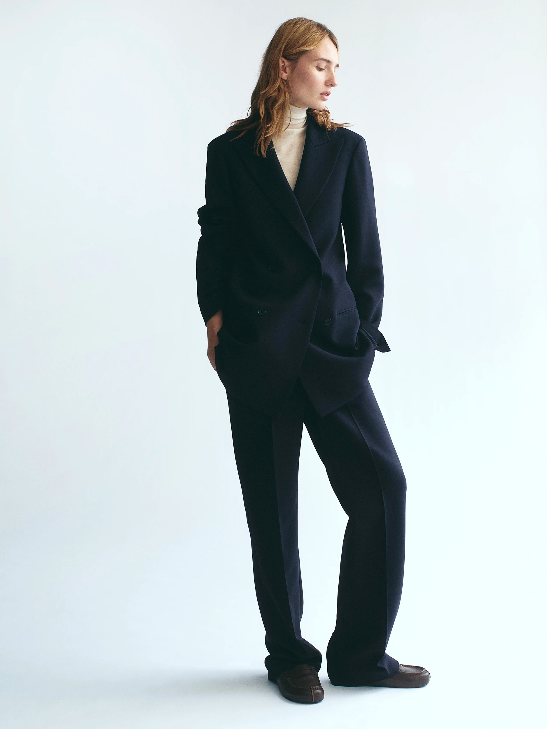 Model standing against a white studio background wearing a navy wool double-breasted wrap blazer with wide lapels, matching wide-leg navy trousers, a cream turtleneck underneath, and dark brown loafers. AW26 D-Line.
