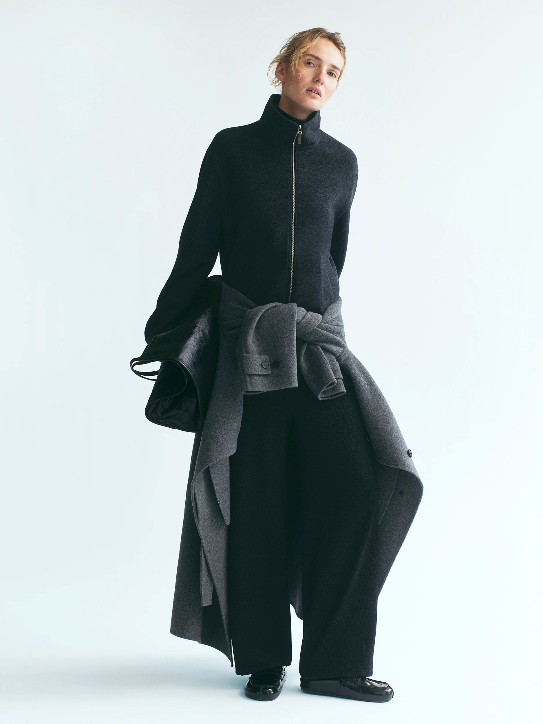 Model standing against a white studio background wearing a fitted black zip-up funnel-neck knit top with flared cuffs and wide-leg black trousers, a dark charcoal wool floor-length coat casually tied around the waist, carrying a black leather drawstring bag. AW26 D-Line.