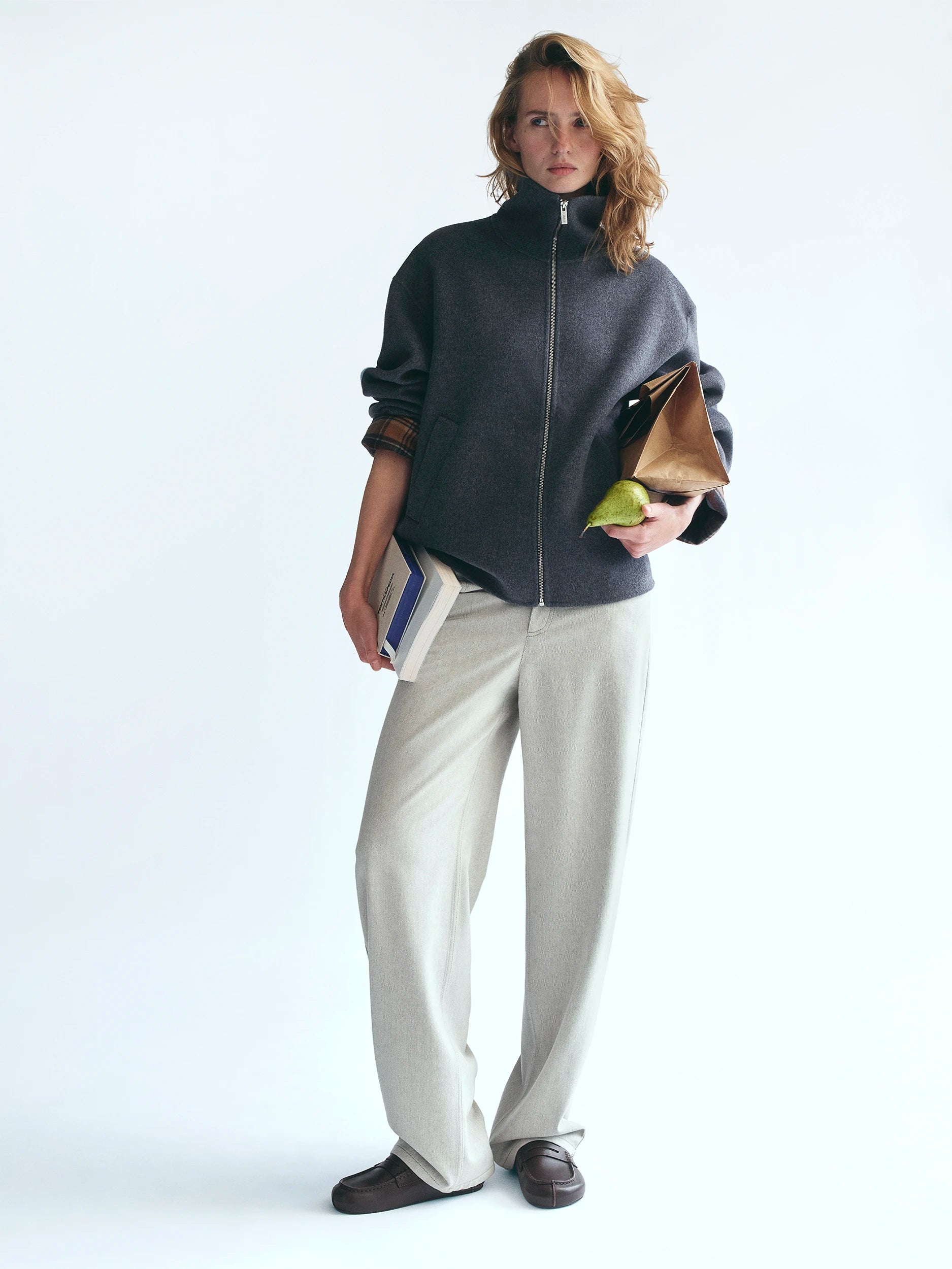 Model standing against a white studio background wearing an oversized dark charcoal zip-up knit jacket with a high funnel neck, paired with wide-leg cream tailored trousers and dark brown loafers, holding a book and a pear. AW26 D-Line.