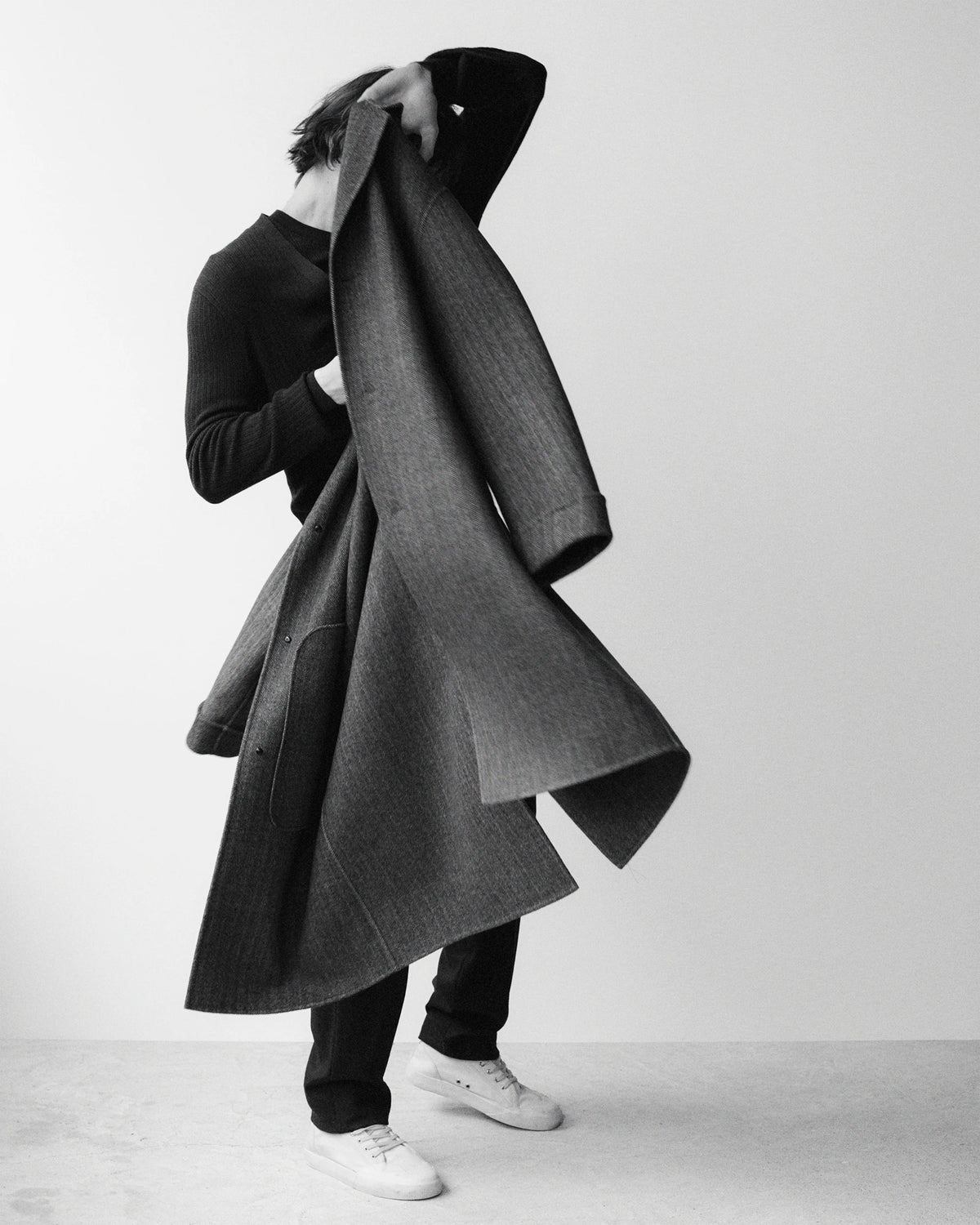 Black and white photograph of a male model lifting a voluminous floor-length dark grey textured wool coat over his head against a white background, obscuring his face, revealing a black crewneck underneath with black slim trousers and white leather sneakers, the coat's dramatic A-line silhouette fully displayed. AW26 Icicle Menswear.
