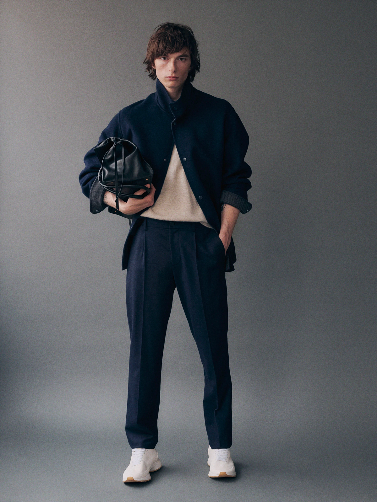 Male model standing against a dark grey background, wearing a navy wool snap-button bomber jacket over a cream crewneck sweater, layered over a dark navy shirt, paired with navy pleated tapered trousers and white chunky sneakers, carrying a black leather drawstring bucket bag. AW26 Icicle Menswear.
