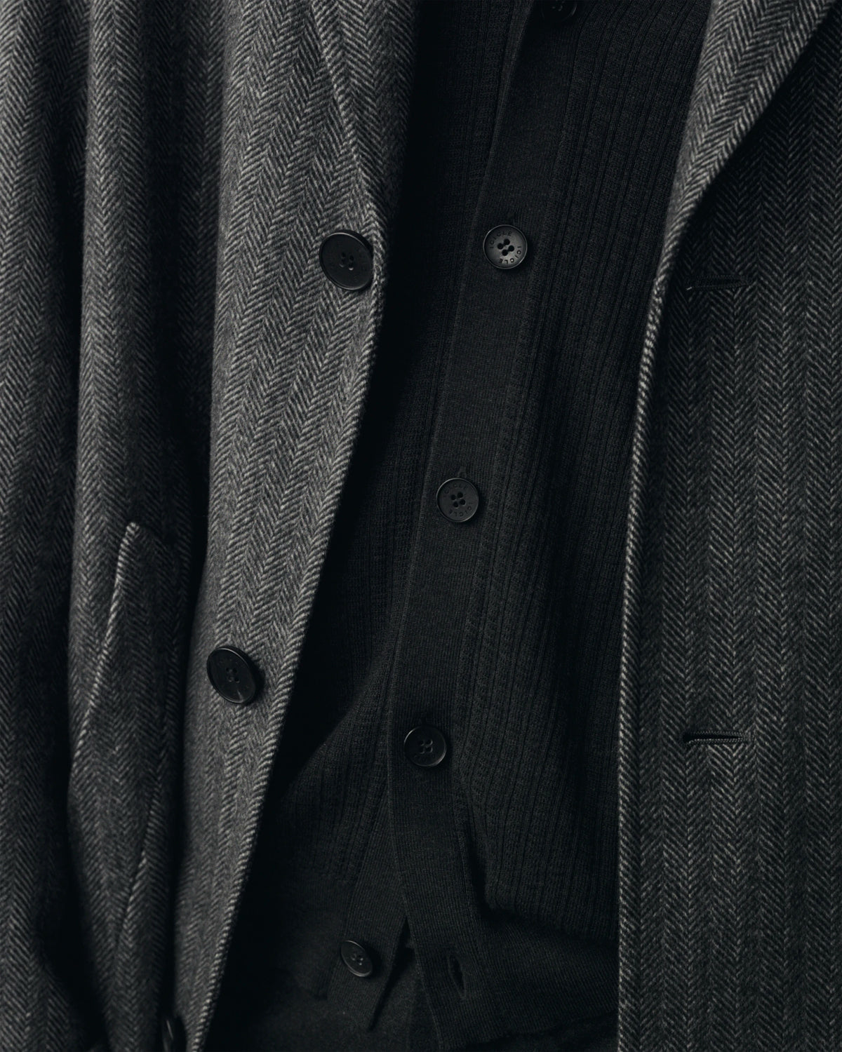 Close-up fabric detail of layered menswear pieces: an open dark grey herringbone wool coat with black horn buttons and a welt pocket, revealing a black ribbed button-front cardigan underneath, with a dark pinstripe lapel visible on the right. AW26 Icicle Menswear.