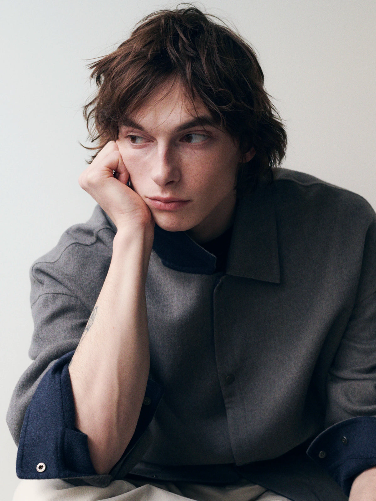 Close-up portrait of a young male model resting his cheek on his hand against a pale grey background, wearing a dark charcoal collarless wool overshirt layered over a navy blue shirt with eyelet cuff detail, with cream trousers visible below. AW26 Icicle Menswear.
