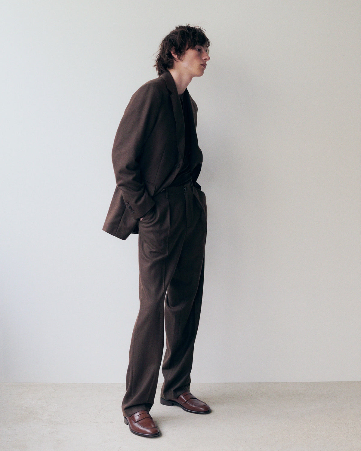 Male model in profile against a white wall, wearing an oversized dark chocolate brown wool blazer with wide lapels and a relaxed drape, layered over a black collarless top, paired with matching wide-leg pleated dark brown trousers and dark burgundy leather penny loafers. AW26 Icicle Menswear.