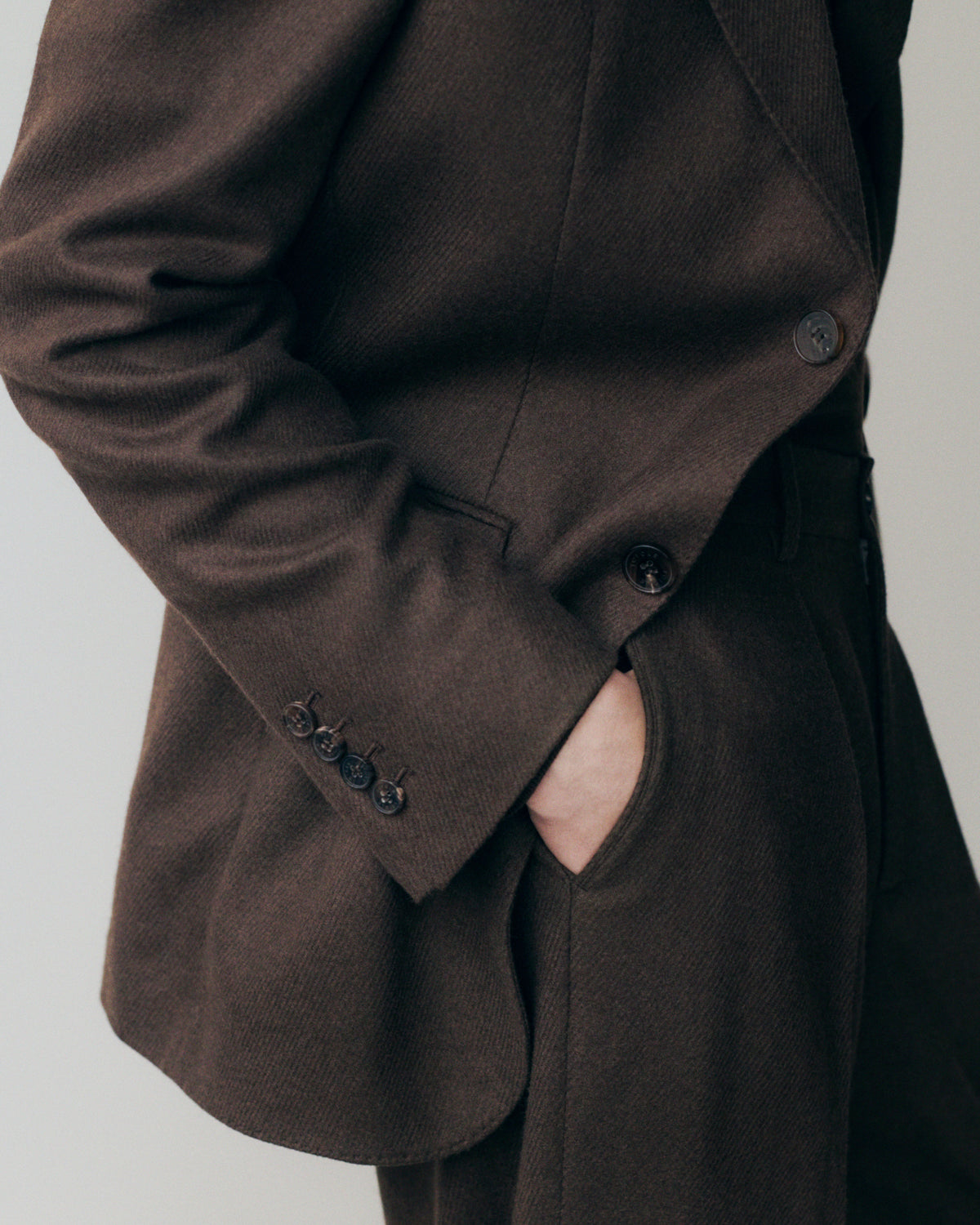 Close-up detail of a dark chocolate brown wool blazer, showing the hand in a side welt pocket, four-button cuff with dark horn buttons, a single-button chest detail, and the fine herringbone texture of the fabric. AW26 Icicle Menswear.