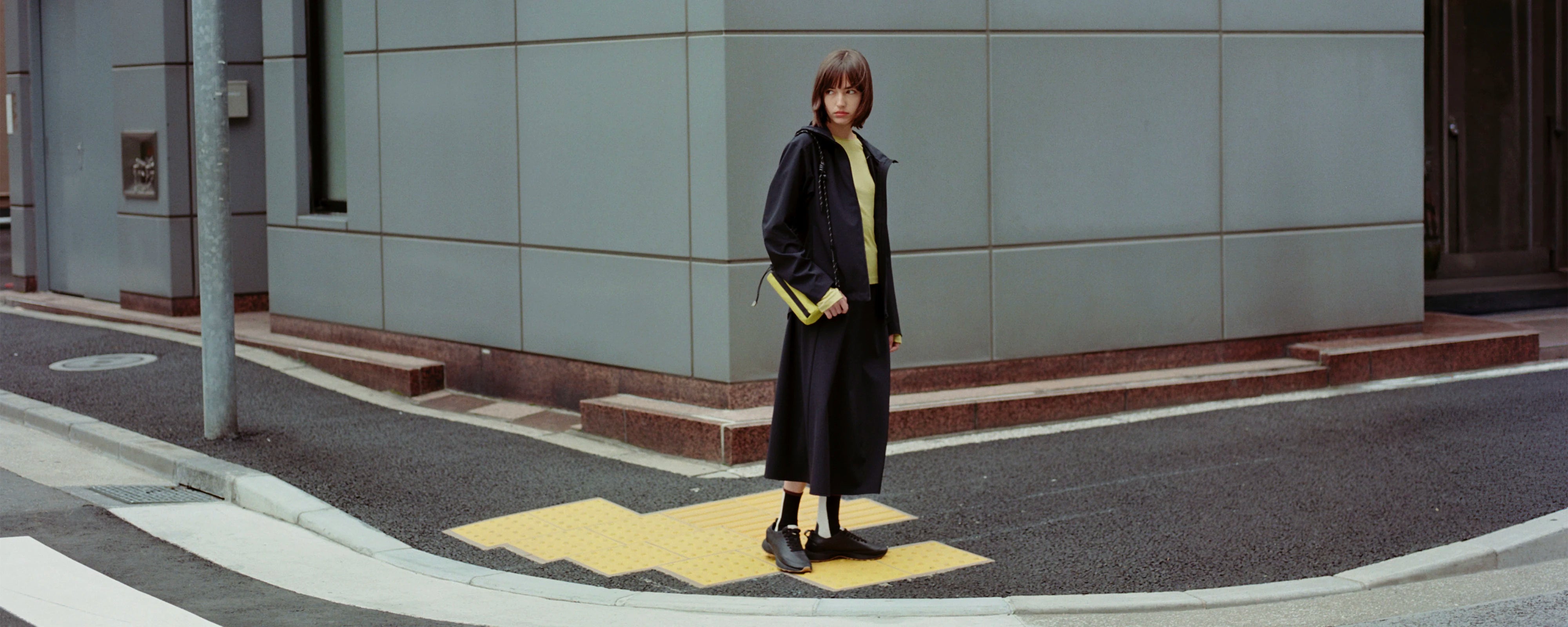 A model stands on a yellow crosswalk at an urban street corner, wearing a long black coat over a yellow top and holding a black bag with yellow trim.
