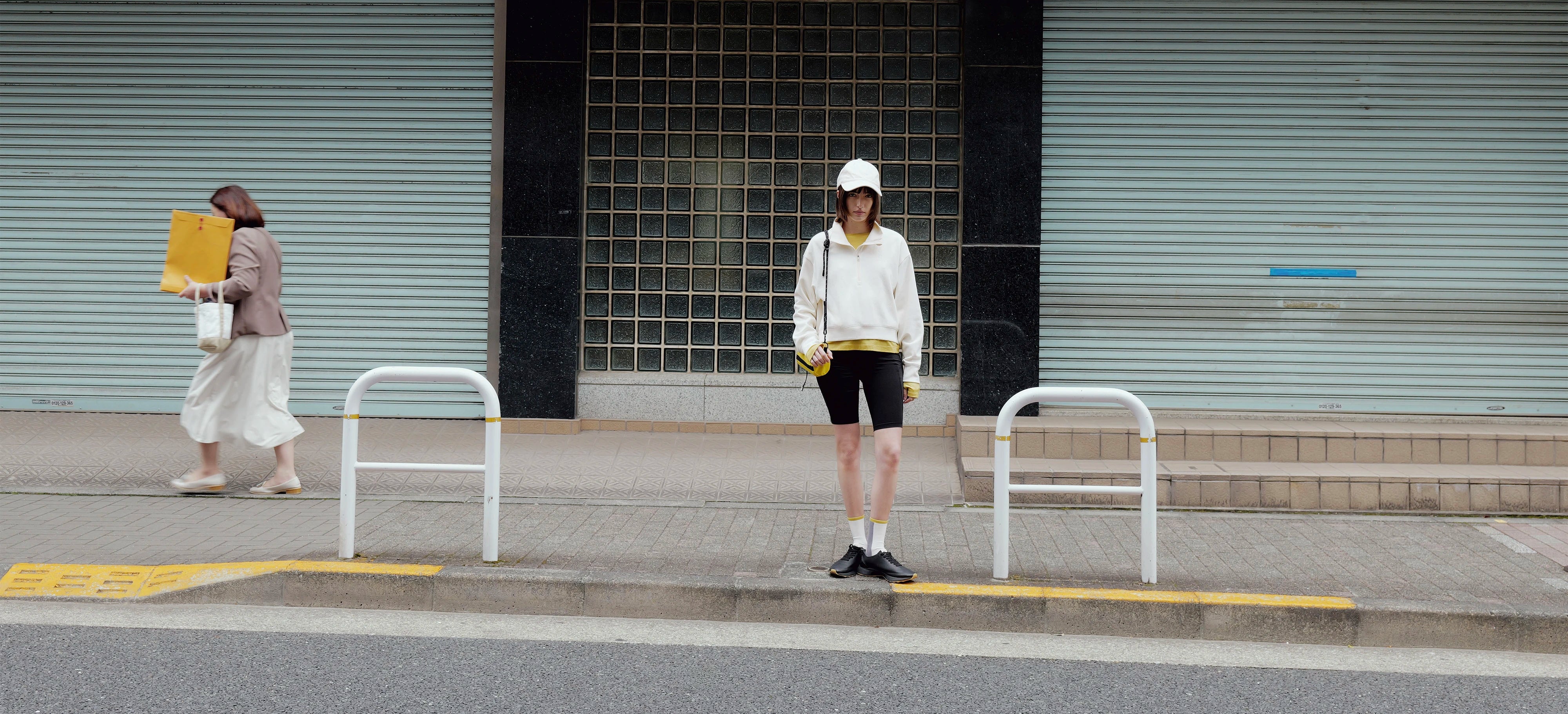 A person wearing a white hoodie, black shorts, and a white cap stands on a sidewalk in front of closed metal storefront shutters. Another person walks past in the background carrying a yellow bag.