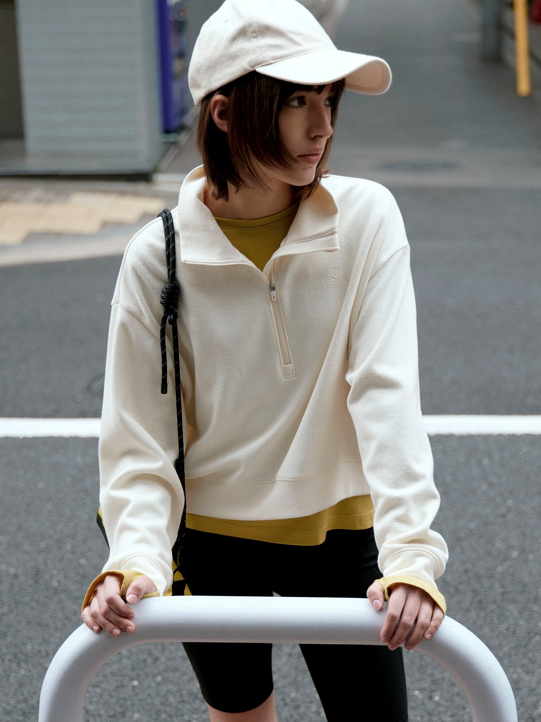 A woman wearing a cream-colored quarter-zip pullover, a matching cream cap, and black bike shorts, holding a white metal barrier on an outdoor street.
