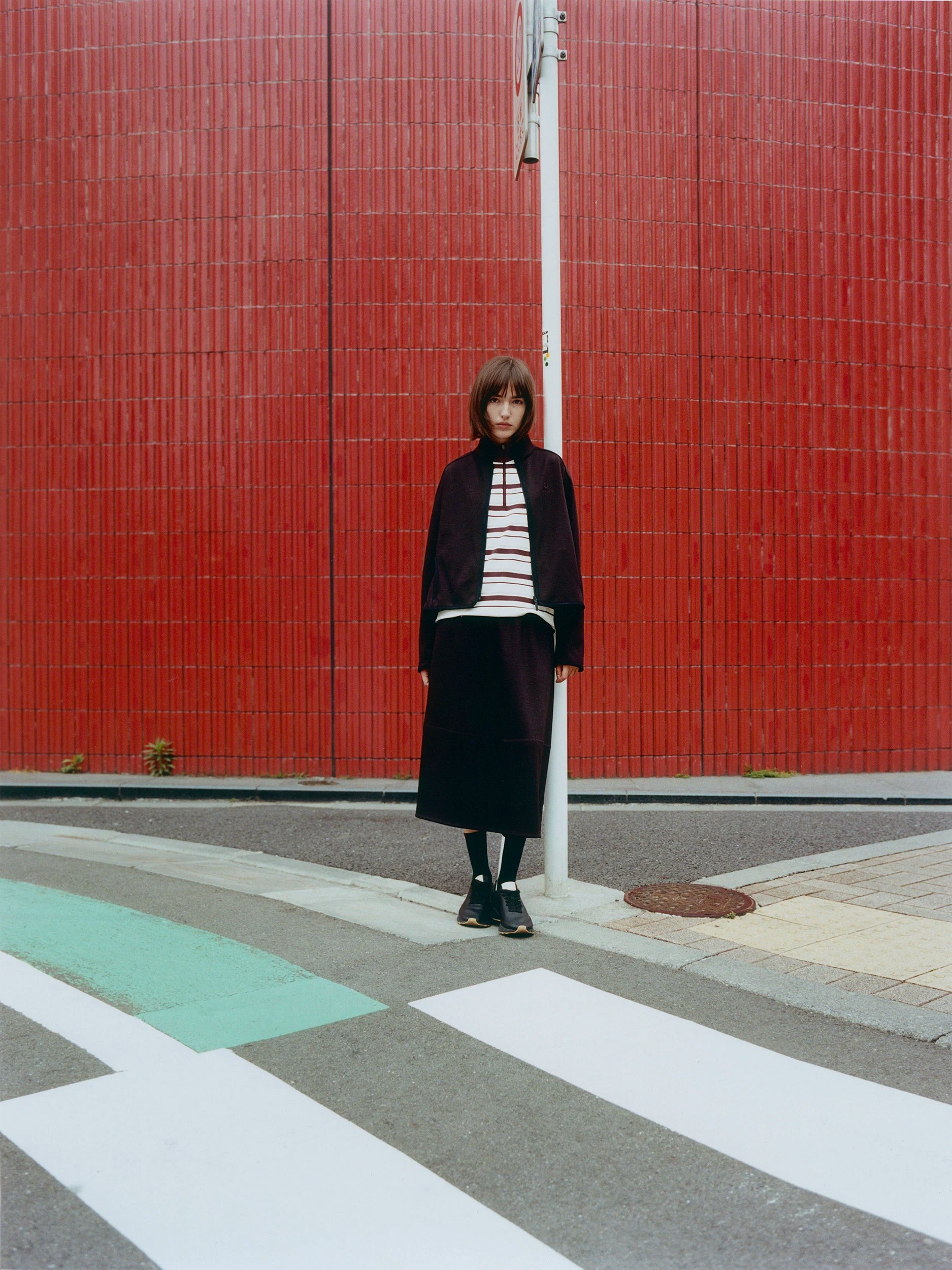 A person stands on a street crosswalk in front of a large red tiled wall, wearing a dark long-sleeve jacket over a white and red striped top with a dark skirt.