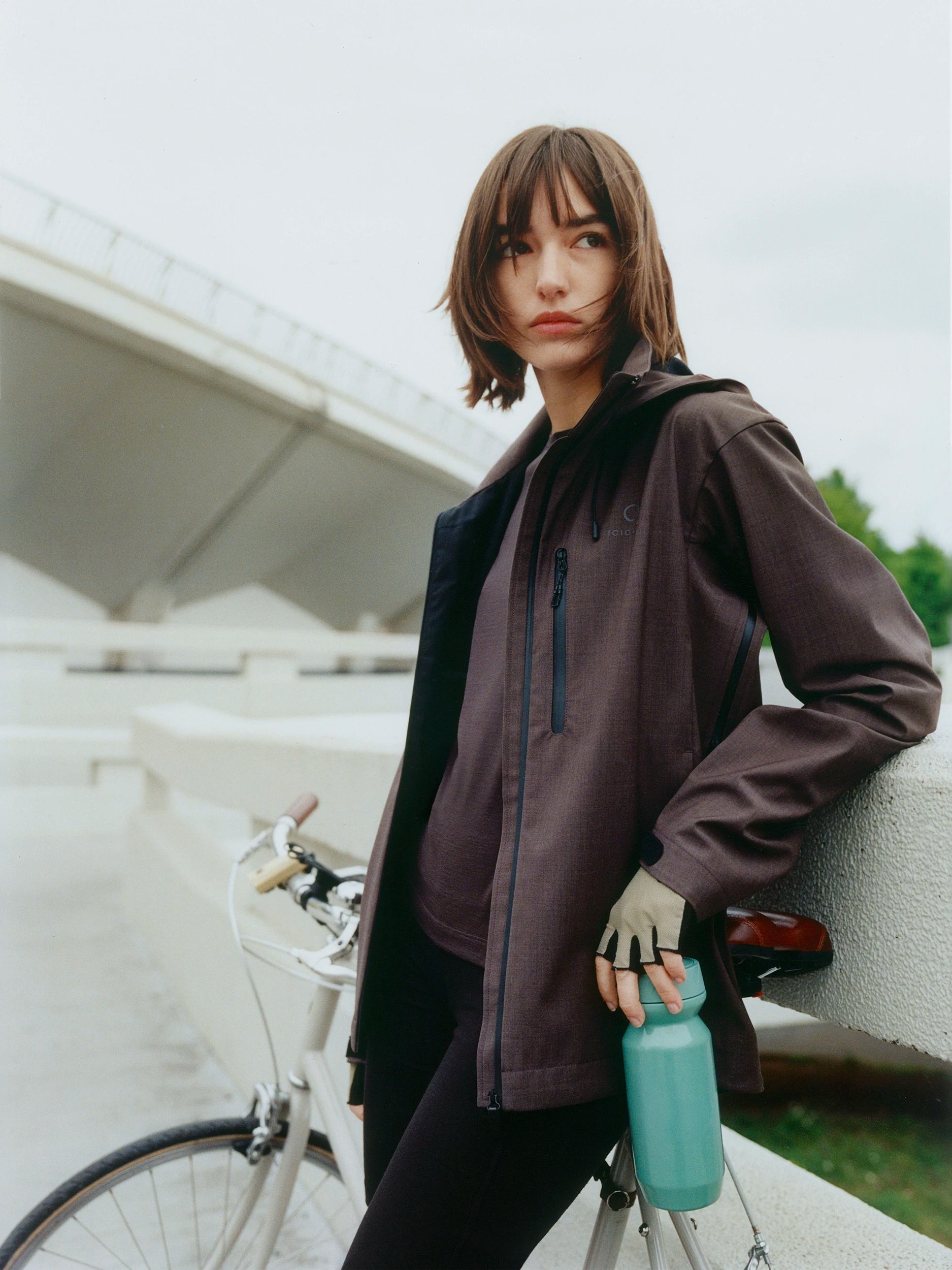 A woman wearing a dark brown hooded jacket and black pants leans against a concrete bridge railing next to a white bicycle, holding a mint green water bottle.