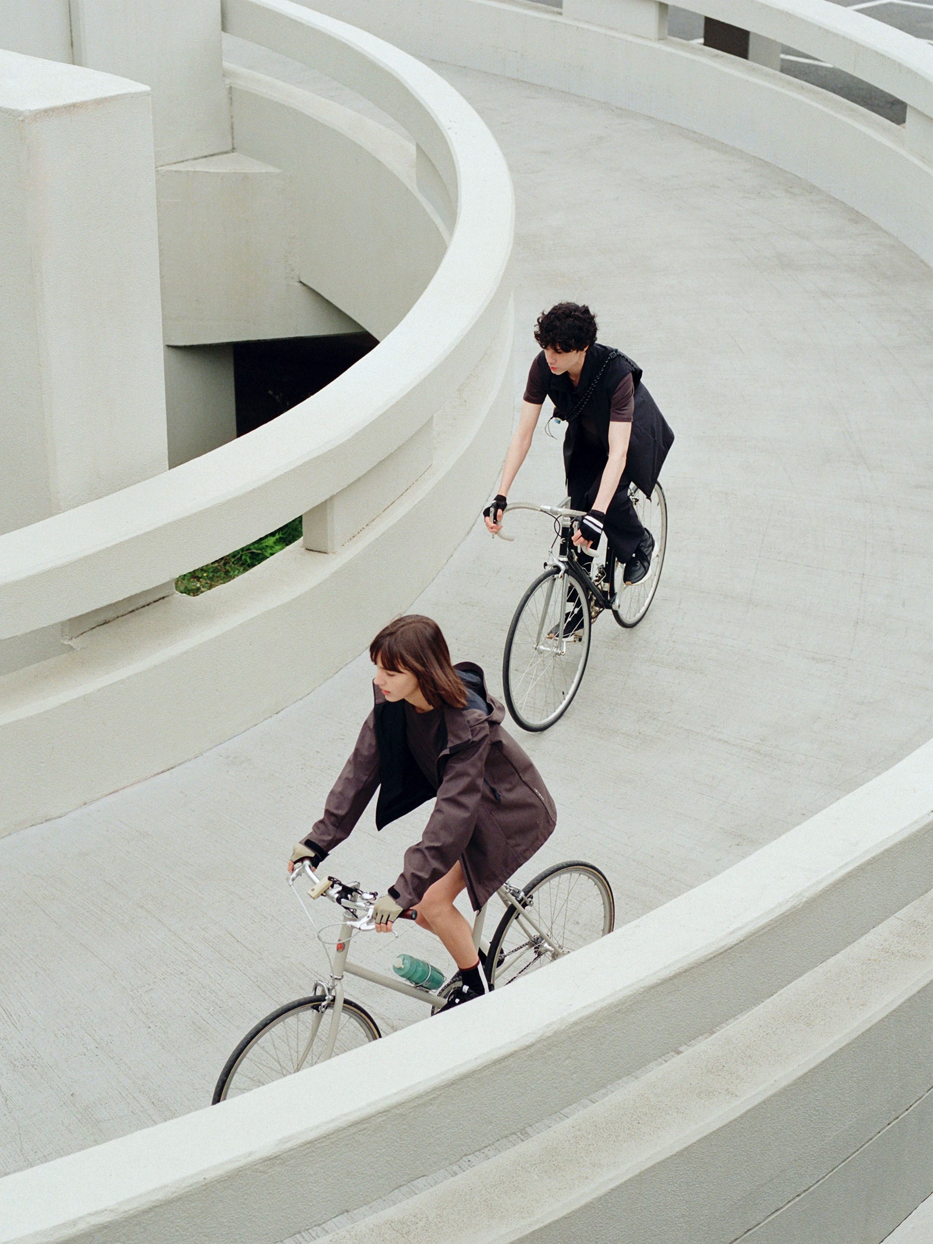 Two people riding bicycles along a curved concrete walkway in a minimalist architectural setting.