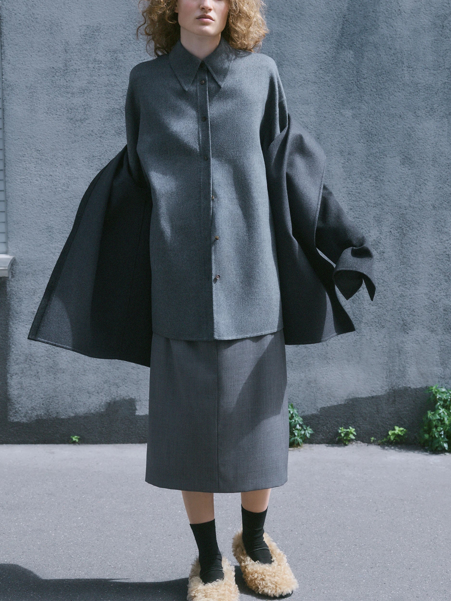 Model standing on a sunlit pavement against a grey concrete wall, wearing an oversized dark grey textured wool shirt-jacket with dramatic wide flared sleeves tied at the cuffs, buttoned front and pointed collar, paired with a matching grey midi pencil skirt, black socks and cream shearling mules. AW26 Icicle Outerwear.