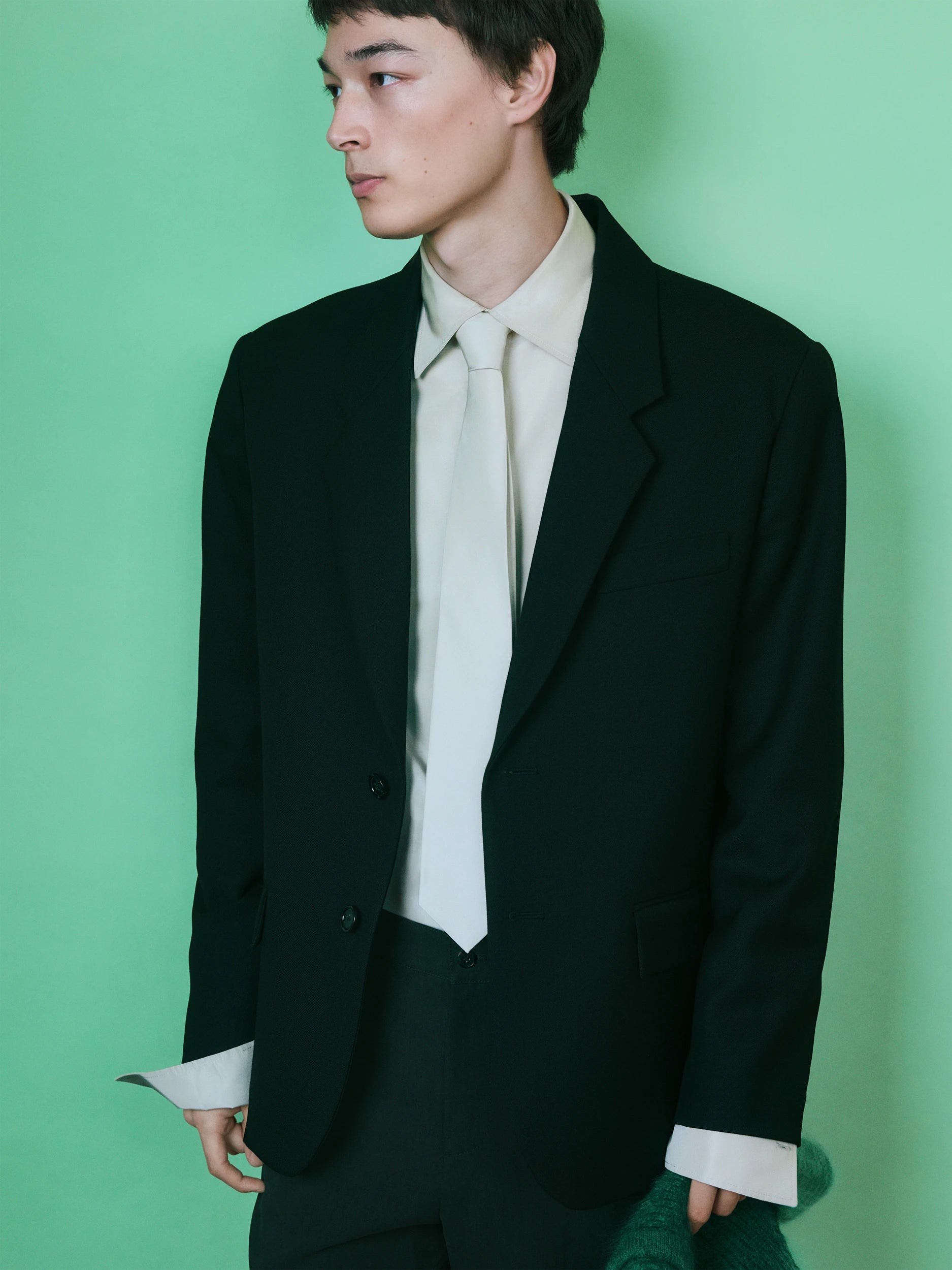 Male model in profile against a bright green background, wearing an oversized dark forest green single-button blazer with wide notch lapels, layered over a cream collarless shirt with a matching cream tie and wide cuffs, paired with dark green trousers. AW26 Icicle Outerwear.