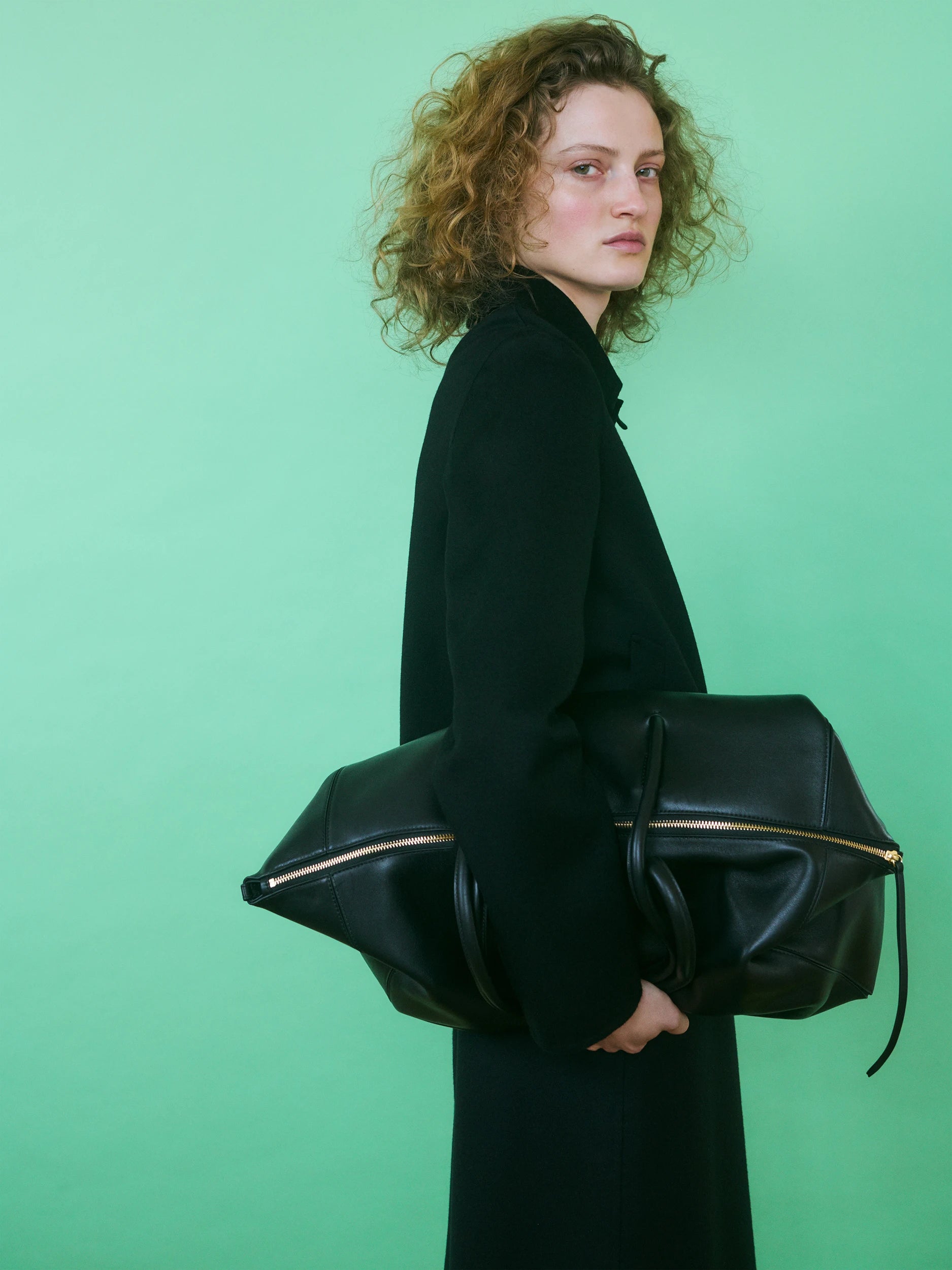 Model in profile against a bright green background, wearing a floor-length black wool coat with a high draped collar, carrying an oversized structured black leather duffle bag with gold zip closures on both sides. AW26 Icicle Outerwear.
