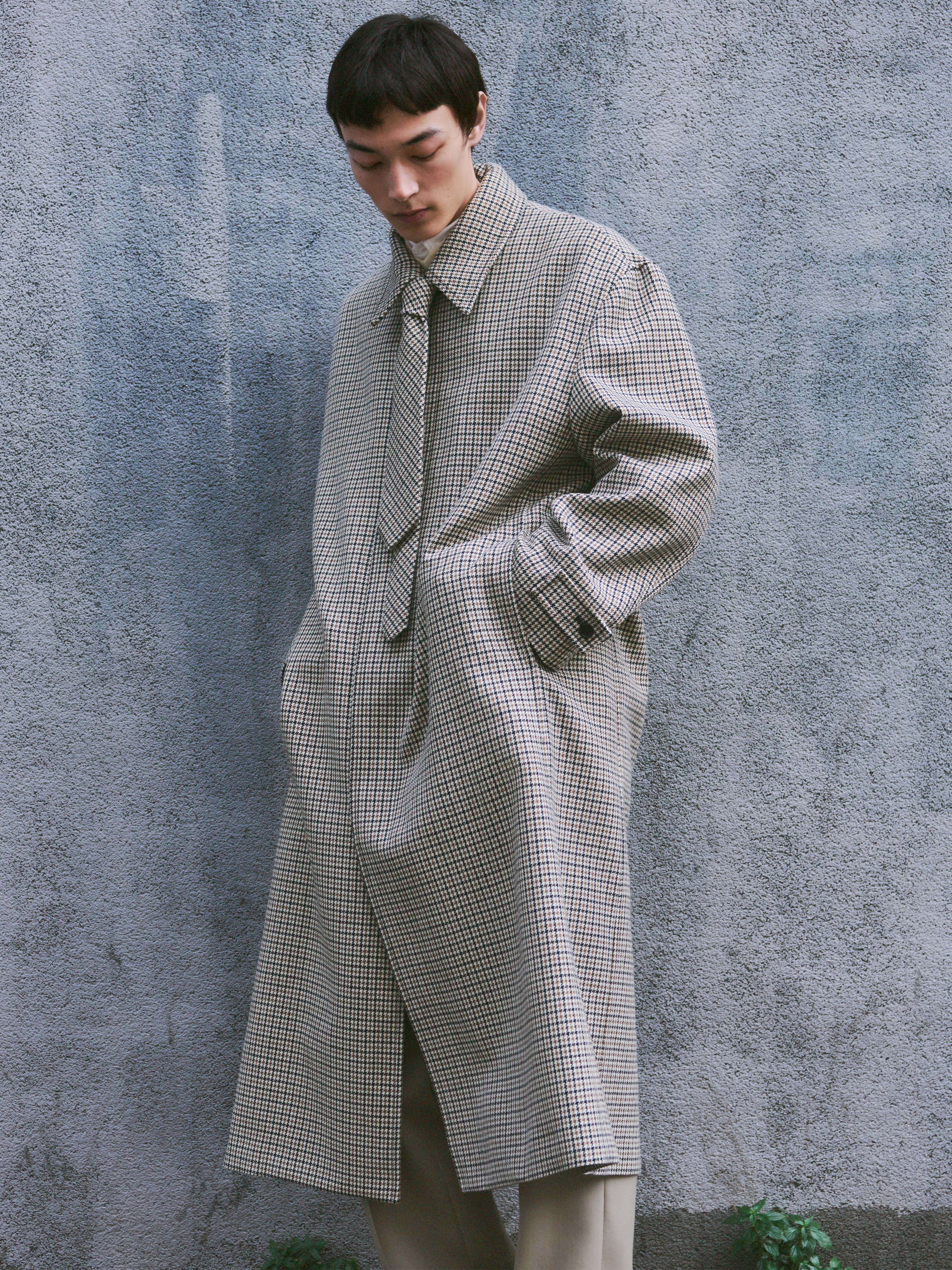 Male model looking down against a textured grey concrete wall, wearing a floor-length oversized houndstooth check wool coat in beige, black and cream, with a wide pointed collar, self-fabric tie belt, front welt pockets and a back vent, paired with cream wide-leg trousers. AW26 Icicle Outerwear.