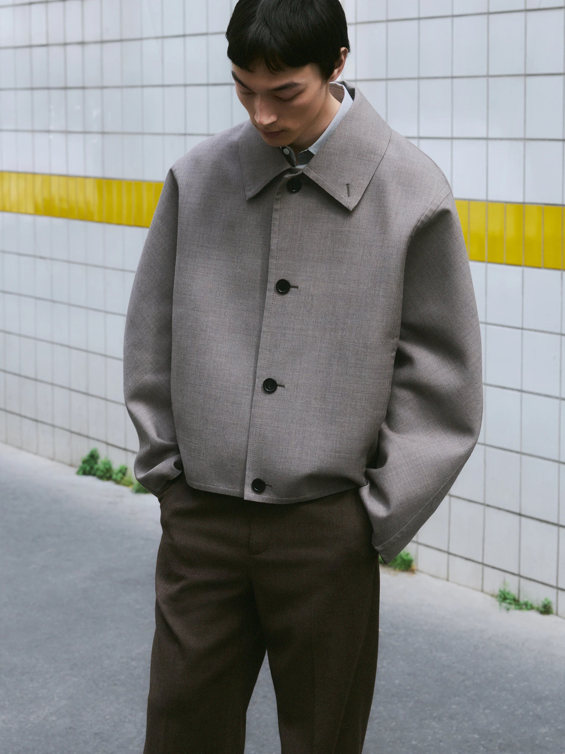 Male model looking down in front of a white tiled wall with a yellow stripe, wearing an oversized boxy grey wool jacket with a wide pointed collar and four black buttons, hands in pockets, paired with wide-leg dark olive brown trousers. AW26 Icicle Outerwear.