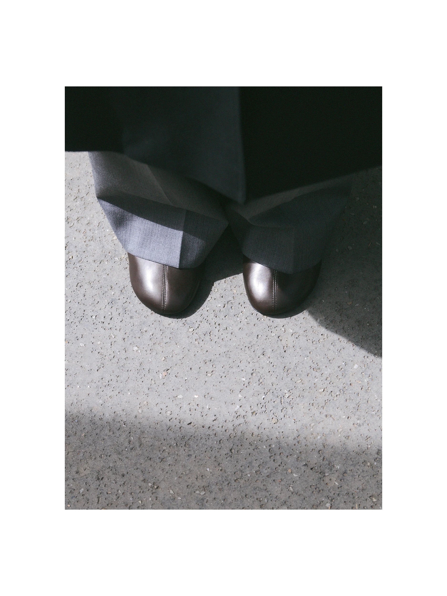 Close-up of a model's feet on a sunlit concrete pavement, wearing a pair of dark gunmetal metallic round-toe ballet flats with a fine seam detail, beneath the hem of wide-leg dark charcoal trousers with a light grey lining visible. AW26 Icicle Outerwear.