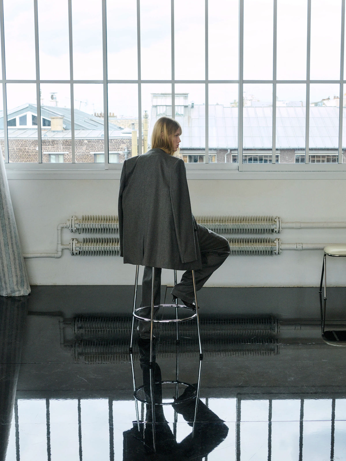 Back view of a female model with blonde hair seated on a tall chrome bar stool, facing large industrial steel-framed windows overlooking Parisian rooftops, wearing an oversized dark grey wool blazer with a centre back vent and matching wide-leg dark grey trousers, reflected in a glossy black floor. AW26 Icicle P-Line.
