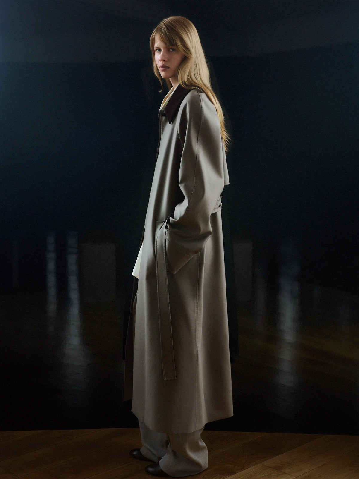 Female model in three-quarter back profile glancing over her shoulder, with long straight blonde hair, wearing a floor-length oversized greige wool coat with a wide dark navy lapel collar, asymmetric open front, side seam pockets and a self-fabric tie belt at the back, paired with matching wide-leg greige wool trousers and black flat mules, against a deep dark teal background with a warm wooden floor. AW26 Icicle P-Line.