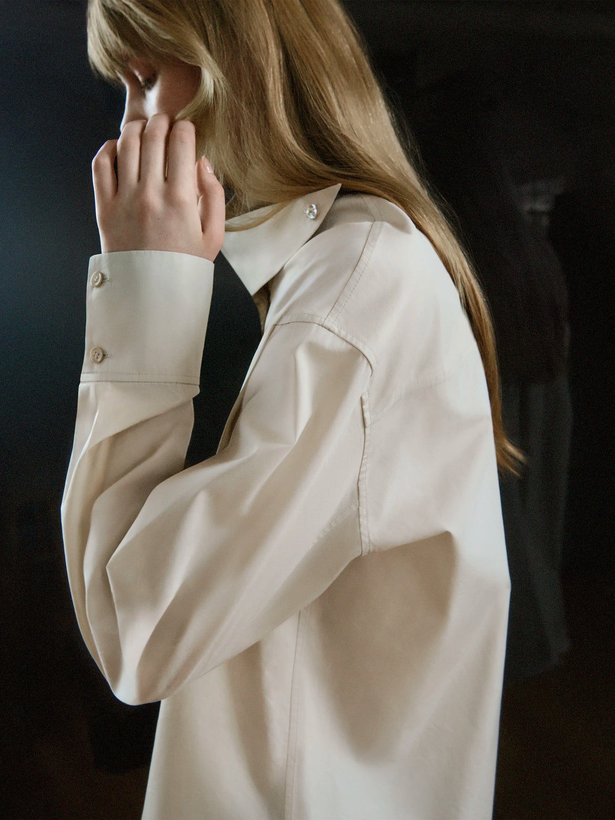 Close-up side profile of a female model with long straight strawberry blonde hair and a fringe, head bowed with hand raised to her face, wearing an oversized sand beige wool shirt-jacket with a wide pointed collar, structured epaulette shoulder seam, voluminous sleeves and a double-button barrel cuff, layered over a black top, against a dark background. AW26 Icicle P-Line.