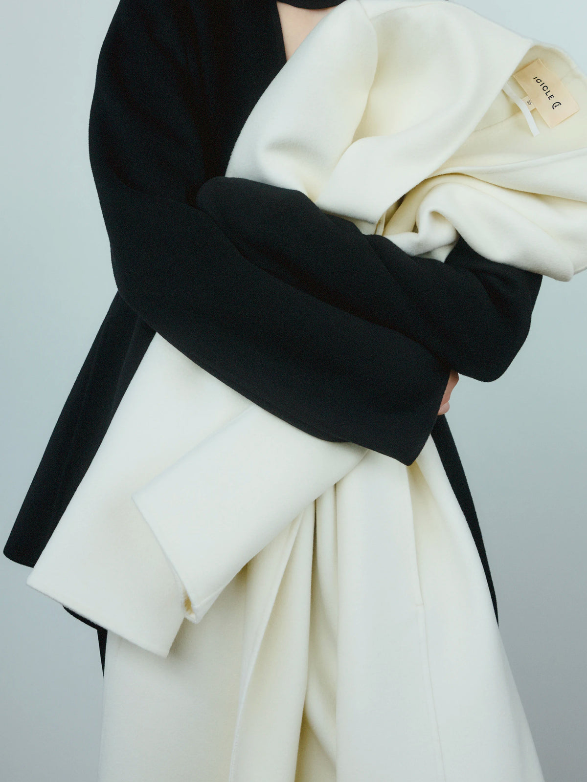 Close-up torso shot of a model wearing a fitted black wool long-sleeve top, arms crossed and cradling a voluminously draped ivory white wool coat with an Icicle woven label visible at the collar, against a pale grey background. AW26 Icicle P-Line.