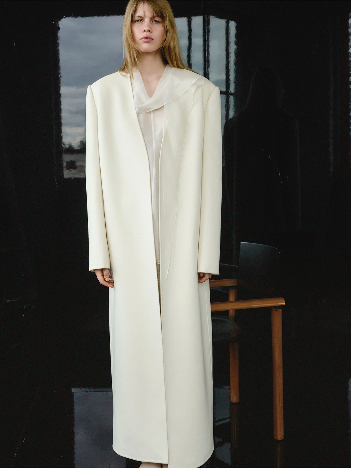 Female model standing in front of tall dark-framed windows with a stormy grey sky beyond, wearing a floor-length oversized collarless ivory white wool coat open over a sheer cream silk cowl-neck slip dress, with a wooden chair visible to the right. AW26 Icicle P-Line.
