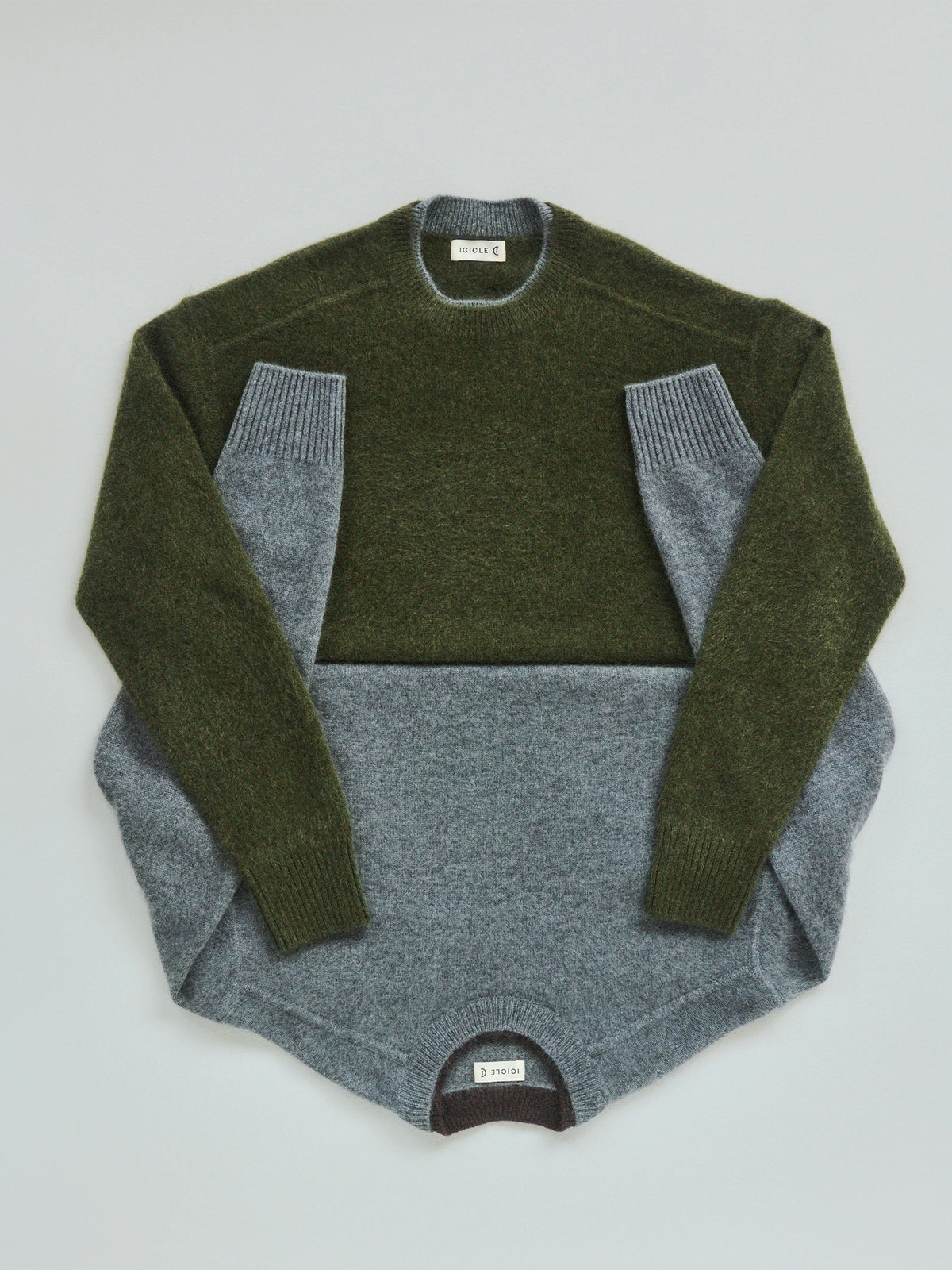 Flat lay of two Icicle wool crewneck sweaters layered together on a pale grey background: a cropped olive green marl knit with ribbed cuffs and collar placed over a longer heather grey marl knit with a curved hem and ribbed hem band, both with woven Icicle labels visible at the neckline. AW26 Icicle P-Line.