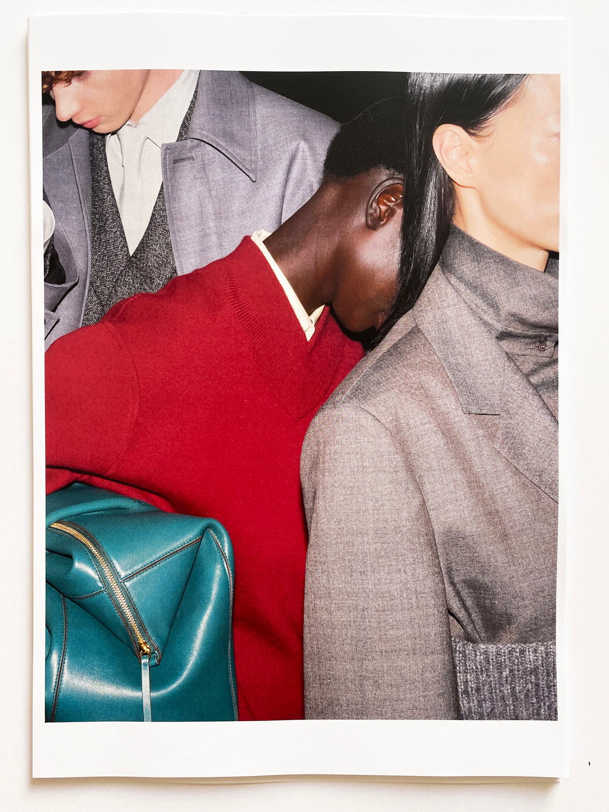 Polaroid-style print showing three models in close proximity: a scarlet red V-neck wool sweater layered over a cream shirt in the centre, paired with a structured teal leather top-handle bag with gold zip detail; a grey tweed blazer with a grey knit visible on the right; and a grey wool jacket with a white shirt and dark tie on the left. AW26 Icicle Atelier Women.