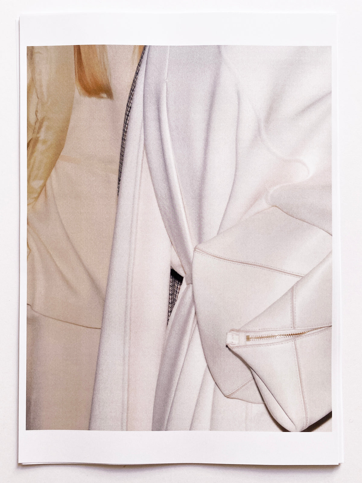 Polaroid-style close-up detail of an ivory quilted silk puffer coat, showing the open front with a fine chain trim along the lapel edge, a structured ivory leather zip pocket at the hem, and a glimpse of a cream knit underneath, with the model's blonde hair visible at the upper left. AW26 Icicle Atelier Women.