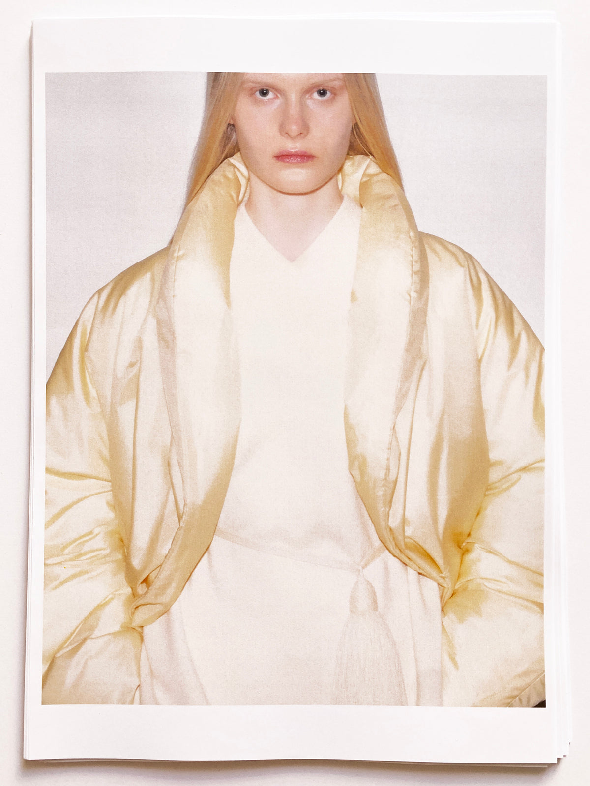 Polaroid-style print of a model with long straight blonde hair, wearing an oversized ivory silk or satin quilted puffer coat with a dramatic draped open collar framing the face, layered over a cream V-neck knit, all-white tonal look, against a white background. AW26 Icicle Atelier Women.