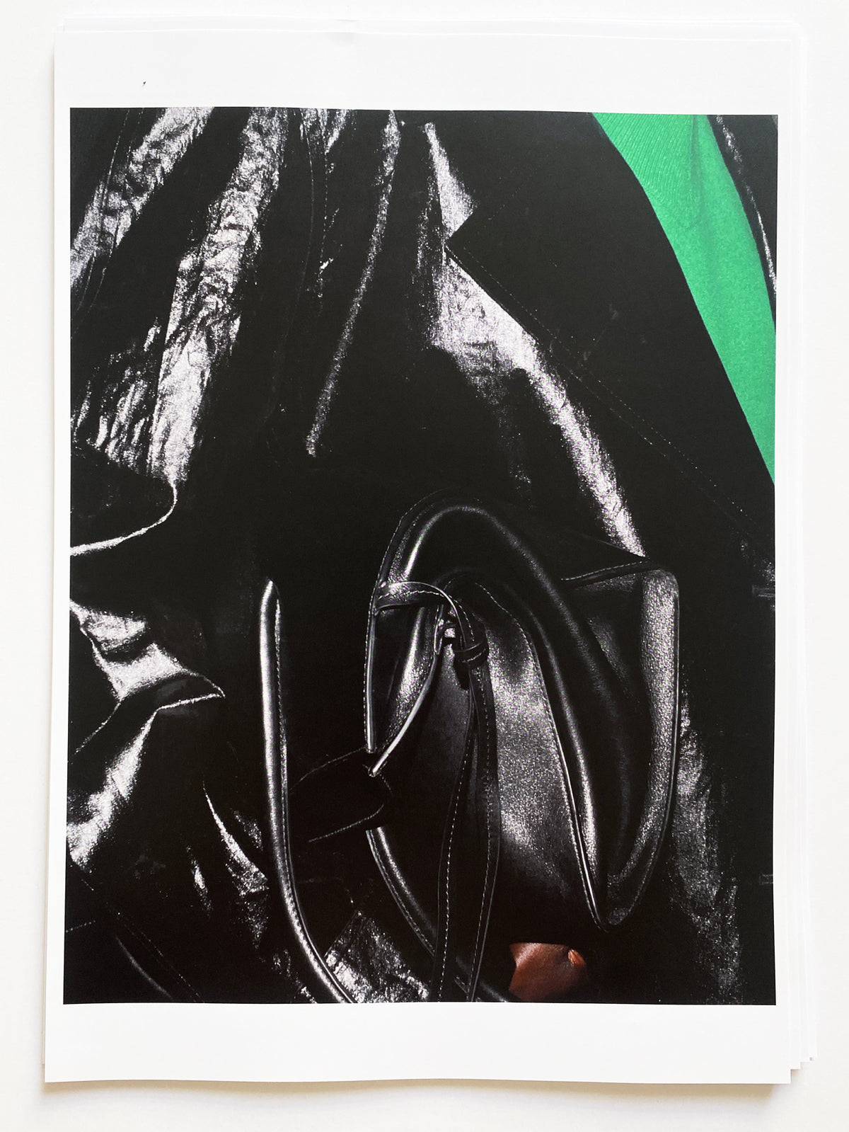 Close-up editorial photograph printed on a white-bordered sheet, showing a structured black leather top-handle bag with double handles and topstitching detail, held against a dramatically draped black glossy fabric with light reflections, with a flash of emerald green leather visible in the upper right corner. AW26 Icicle Atelier Women.