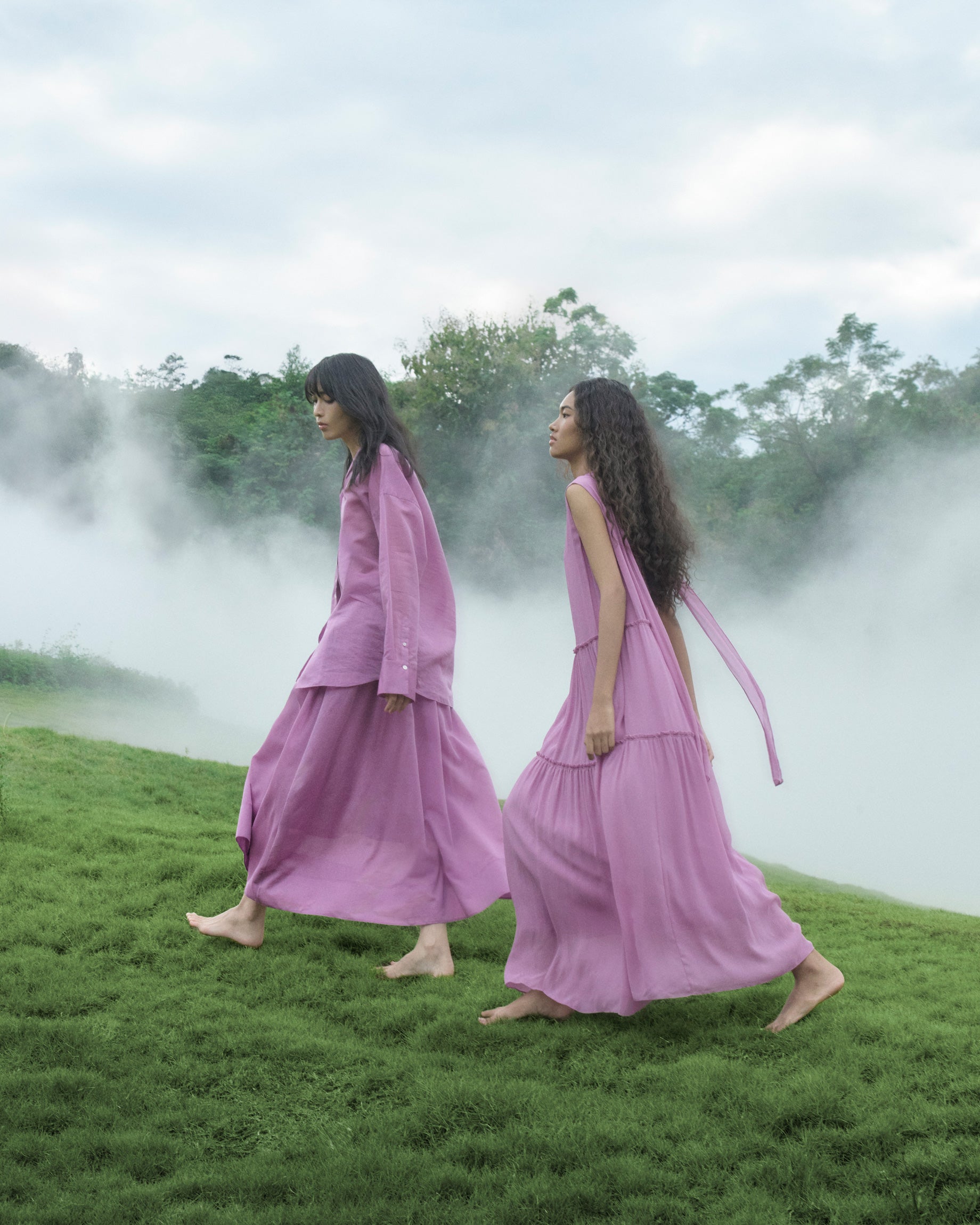 Two women in matching pink outfits standing on a grassy field with mist and trees in the background.